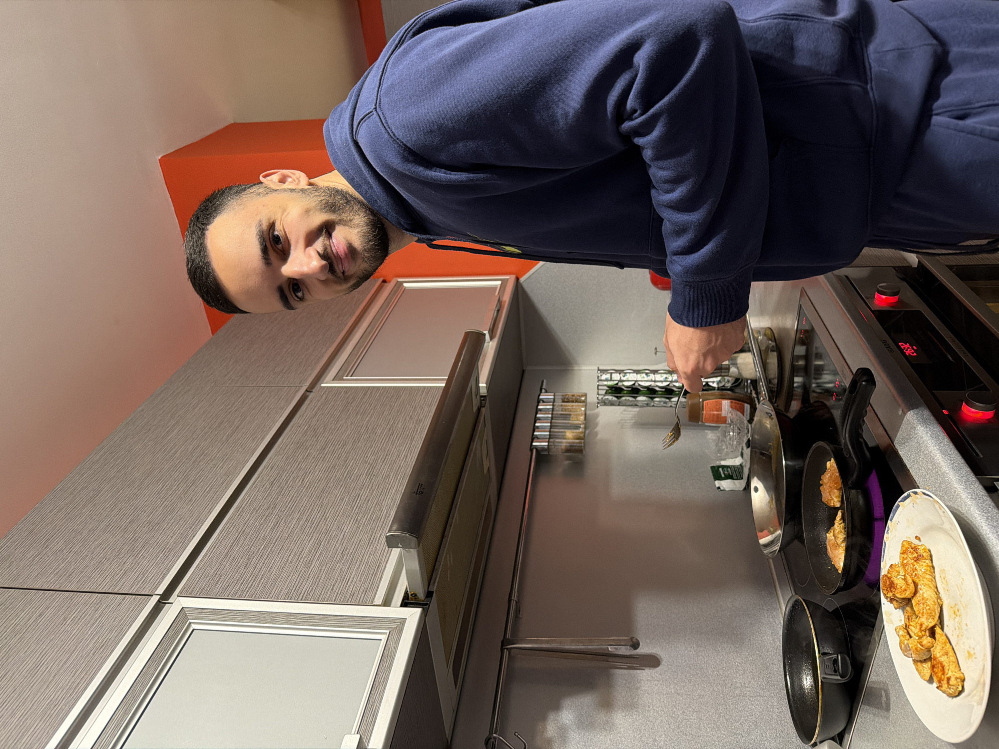 A person with short dark hair and a beard, wearing a dark blue hoodie, is standing in a kitchen. The kitchen has gray cabinets with silver trim, a stainless steel countertop, and a black stovetop with red controls. Two black frying pans are on the stovetop, one containing food being cooked. Cooked food is also present on a white plate with a blue and gold rim. A fork is visible in the person's hand, near a collection of spices and cooking oils. The background includes a wall painted orange near the ceiling. The image does not contain enough information to determine the exact location in Plovdiv, Bulgaria.