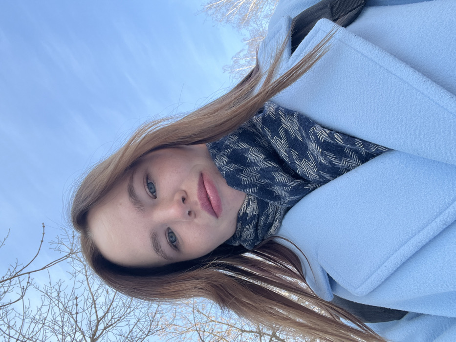 A young woman with light brown hair, fair skin, and blue eyes poses in a tilted selfie. She is wearing a light blue coat and a patterned scarf. The sky is visible in the background, along with bare tree branches. The photo is taken from a low angle.
