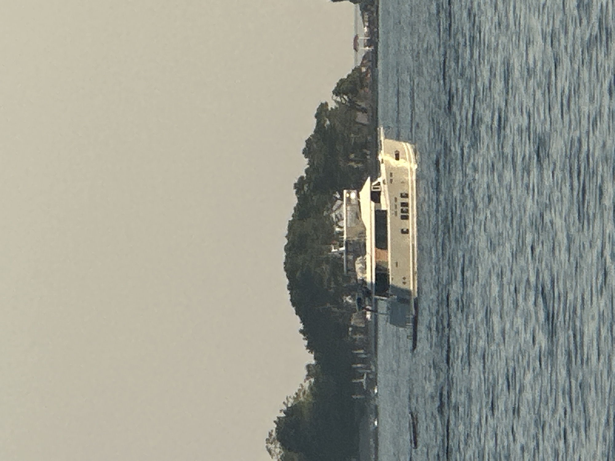 A wide-angle, eye-level outdoor daytime shot captures a light-colored motor yacht navigating calm, choppy blue water. The yacht is positioned in the mid-ground, slightly to the left of the frame's center, and appears to be moving away from the viewer. It has multiple decks, large windows, and what looks like a radar dome or antenna on its upper level. The boat's hull is white with a beige stripe and lettering that is unreadable. In the background, a densely wooded island with a rocky shoreline forms a dark green mass. Scattered along the island's edge are hints of structures, possibly buildings or piers, and a small, white gazebo-like structure is visible on the far right of the island. The sky is a pale, hazy gray, suggesting an overcast or hazy day. The overall scene depicts a tranquil coastal environment, likely near a populated area given the presence of the yacht and background structures. The location is not definitively Budva, Montenegro, as there are no specific landmarks or identifying features to confirm this.