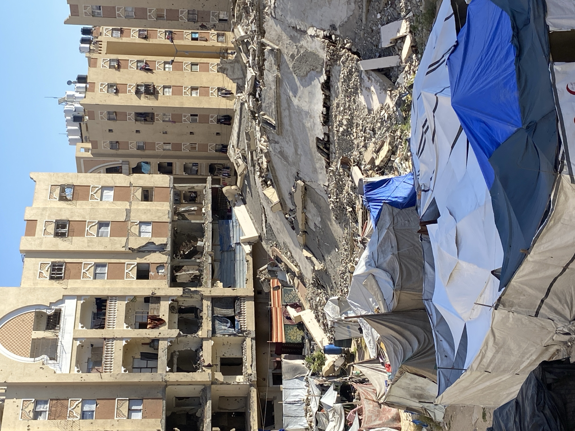 The image displays a scene of widespread urban devastation in Khan Yunis, Palestinian Territory, under a clear, bright blue sky. On the left, a tall, multi-story residential building, painted light brown/beige, stands partially intact but shows significant damage, including shattered windows, missing facade sections, and possible bullet holes. Numerous white water tanks are visible on its roof, and balconies contain debris and household items. The building features distinctive white decorative elements around windows and balconies.
Adjacent to this, the central and right portions of the image are dominated by a massive pile of concrete rubble, twisted rebar, and fragmented building materials, indicating the complete collapse of one or more structures.
Amidst this destruction, on the right side, a makeshift encampment of tents and shelters is erected. These shelters are constructed from various colored tarpaulins, including blue, white, gray, and black, some appearing umbrella-shaped while others are more rectangular, suggesting temporary housing for displaced residents.
Within the damaged and exposed rooms of the partially standing buildings, personal belongings like clothes and carpets are visible. On one white tent, a discernible red crescent logo with the text "Palestinian Red Crescent Society" is present, indicating humanitarian presence. The strong sunlight casts sharp shadows, suggesting the time of day is around midday. The overall scene conveys immense destruction, loss, and the resilience of a community adapting to severe displacement.