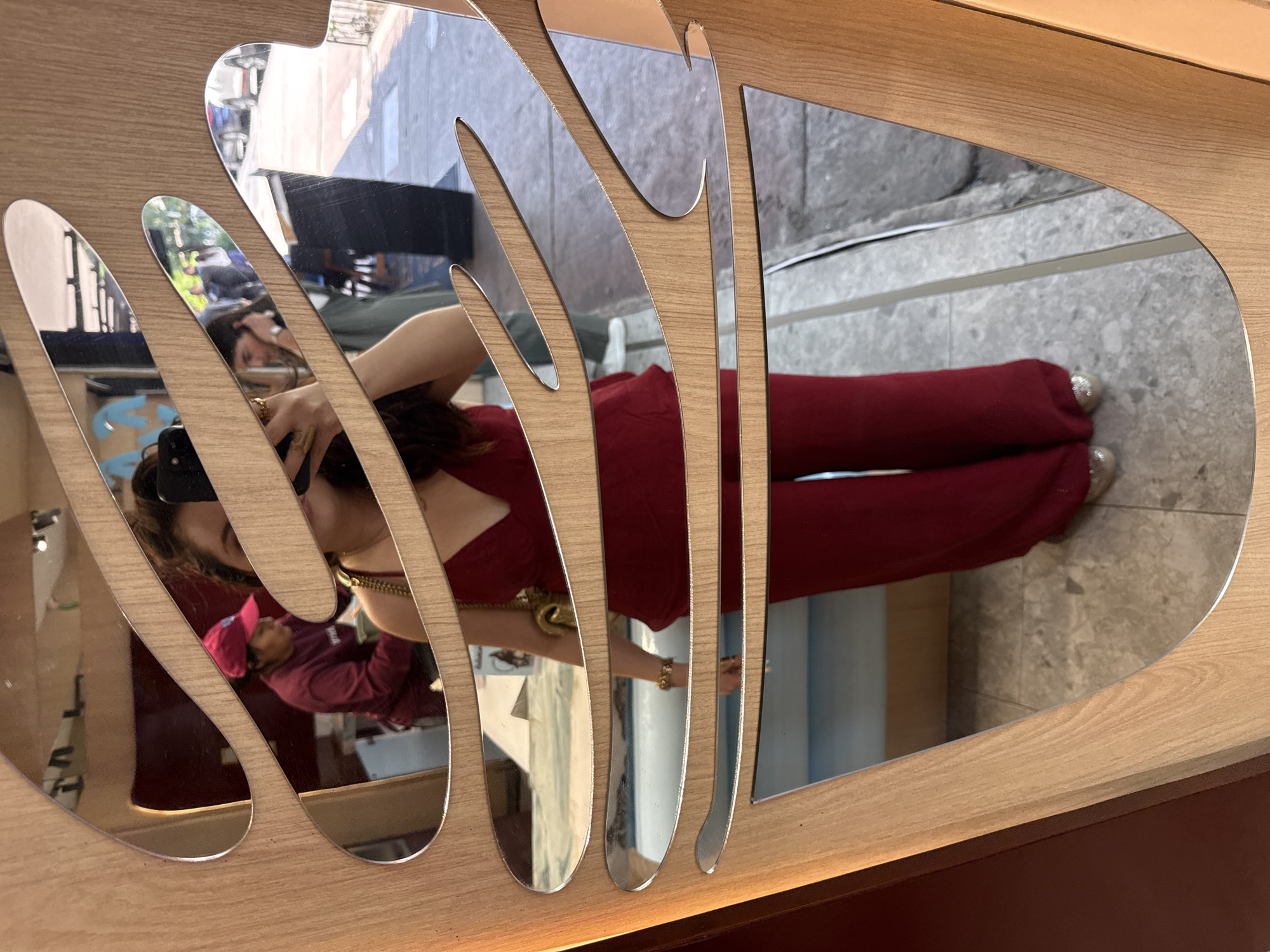 The image depicts a mirrored surface with irregular, organic-shaped cutouts mounted on a wooden panel. Reflected in the mirror is a woman wearing a red jumpsuit and a gold necklace. She holds a black object, likely a phone, in her hand and appears to be taking a picture of herself. The reflections also reveal a glimpse of another person wearing a pink hat and a maroon jacket. The backdrop includes elements of an urban environment, possibly a sidewalk or street. The location is inferred to be Mexico City, Mexico, though this is not explicitly visible in the image. The lighting is natural, suggesting the scene is illuminated by daylight.