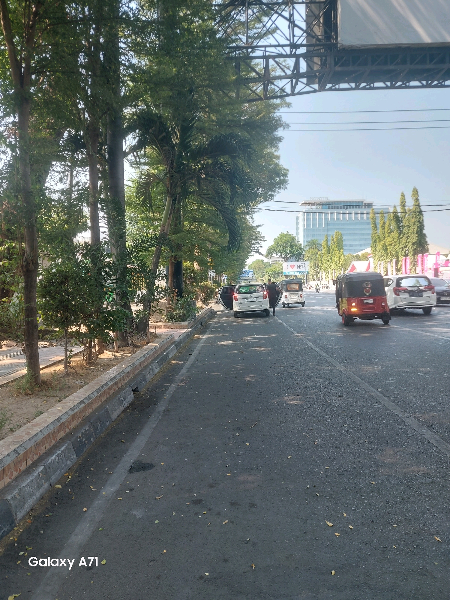 The image depicts a street scene in Makassar, Indonesia, likely taken on a sunny day. The foreground features an asphalt road with a distinct white line marking a lane. A concrete curb with a decorative tiled edge runs along the left side of the road, separating it from a planted area with trees and shrubs.
In the middle ground, several vehicles are visible on the road. A white car with its driver's side door open is parked on the left. Behind it, a white three-wheeled auto-rickshaw, commonly known as an "ojek" or "becak motor," is also parked. Further down the road, a red and yellow three-wheeled auto-rickshaw is in motion, appearing to be a passenger vehicle. To the right of the auto-rickshaw, a white SUV is also on the road.
The background reveals a cityscape with a tall, modern glass building, identified by a sign that reads "NOVOTEL". Green trees line the street, and a large overhead structure, possibly a billboard or a bridge, is visible in the upper portion of the image. Power lines are strung across the sky. The overall impression is of a busy urban environment with moderate traffic.
