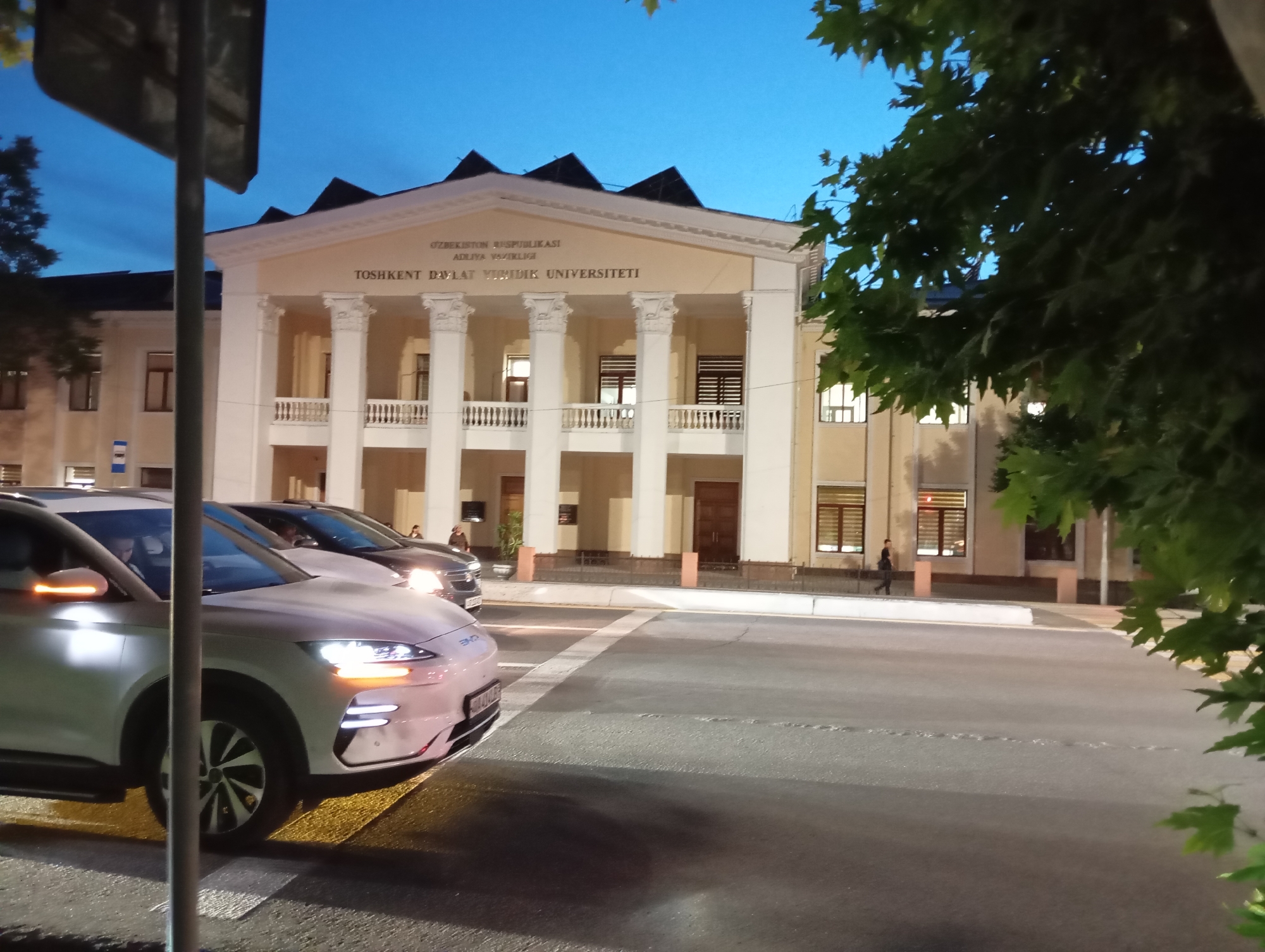 The image shows a street scene in Tashkent, Uzbekistan at dusk. The building in the background is the Tashkent State University of Law, identified by the sign above its columned portico. Several cars, including a white electric vehicle with distinctive headlights, are stopped at a crosswalk in the foreground. A pedestrian is visible walking along the sidewalk near the university building. Trees are present on the right side of the frame.