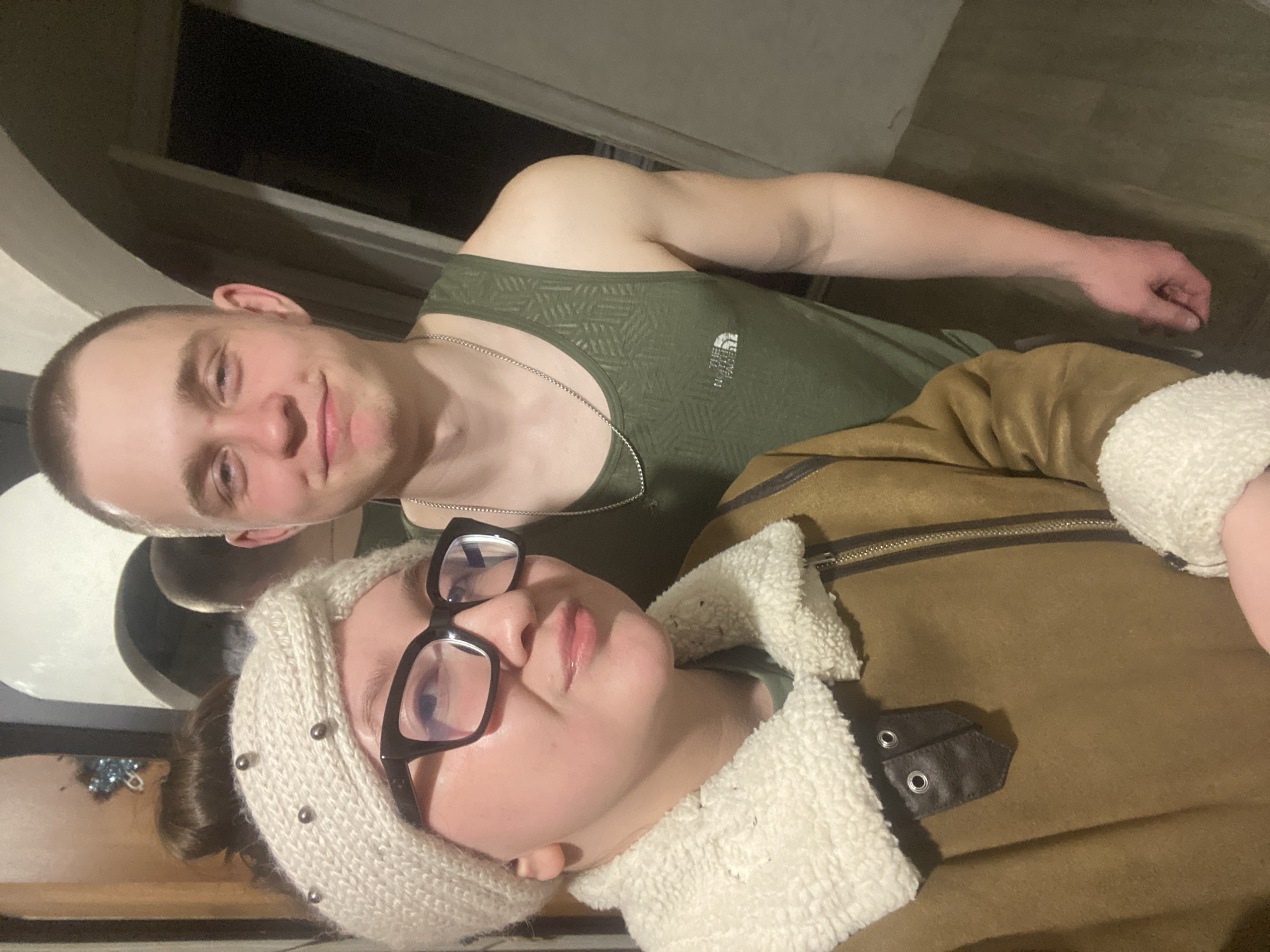 The image displays two individuals indoors, likely in a residential setting within Chaikovsky, Russia, though the specific location is not discernible. In the foreground on the left, a light-skinned female individual is positioned, wearing a cream-colored knitted headband adorned with silver studs, black-framed glasses, and a brown faux shearling jacket with white fleece lining on the collar and cuffs. Her hair is styled in a bun, and she is looking towards the camera with pursed lips. Behind her, to the right, stands a light-skinned male individual with short, buzz-cut hair. He is wearing a dark green tank top with a subtle geometric pattern and a "The North Face" logo on the chest, along with a thin silver chain necklace. He is looking directly at the camera with a faint smile. A reflection of the male individual's head is visible in a mirror or reflective surface behind the female individual. The background features a light-colored arched opening and a dark vertical recess or doorway. A bluish, possibly decorative object is partially visible in the upper left corner of the background. The floor appears to be a light-colored wood-look laminate or vinyl.