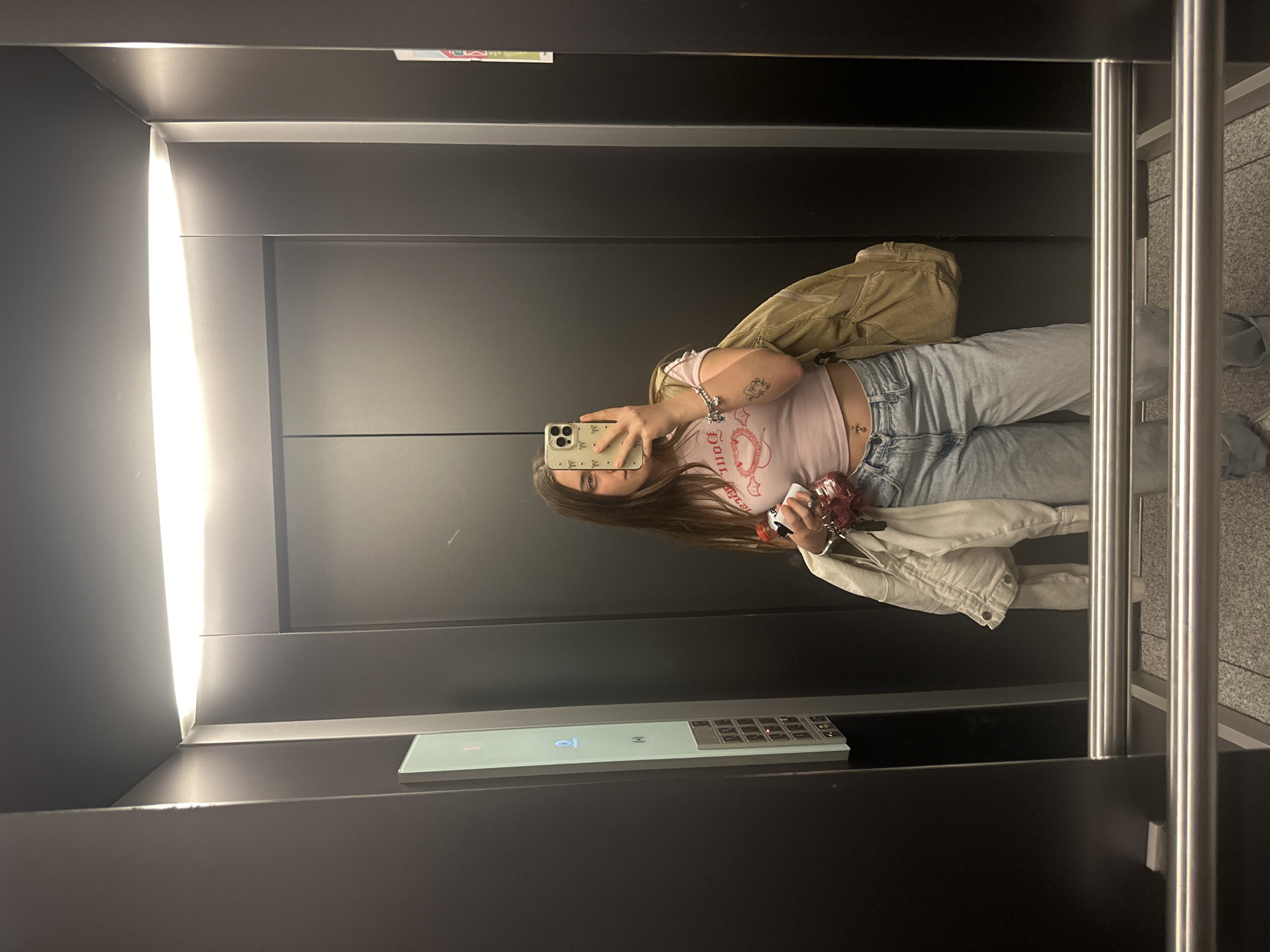 A young woman is depicted taking a mirror selfie inside a modern elevator, located in Huixquilucan de Degollado, Mexico. The image is oriented sideways, with the woman and elevator appearing rotated 90 degrees clockwise.
The subject is a young woman, likely in her late teens or early twenties, with long brown hair. She is holding a light-colored smartphone with a patterned case, using it to capture her reflection. She is wearing a light pink crop top with red text, possibly reading "Dolls," light-wash blue jeans, and a light beige or off-white jacket, possibly corduroy, draped over her arm. Accessories include bracelets, rings, and a stomach piercing. A small, stylized tattoo is visible on her left arm. She also carries a beige bag or backpack over her shoulder.
The elevator interior features dark, reflective wall panels and a bright vertical light strip illuminating the left side of the space. On the right, there are sleek metal handrails and a section of the elevator floor, which appears to be speckled grey. A modern elevator control panel is visible on the right wall, featuring a glowing digital screen that displays a "P" or "1" (likely indicating a floor number) and an array of illuminated buttons and a keypad. The time of day and weather are not discernible, as the scene is indoors with artificial lighting. The overall aesthetic of the elevator is clean and contemporary.
