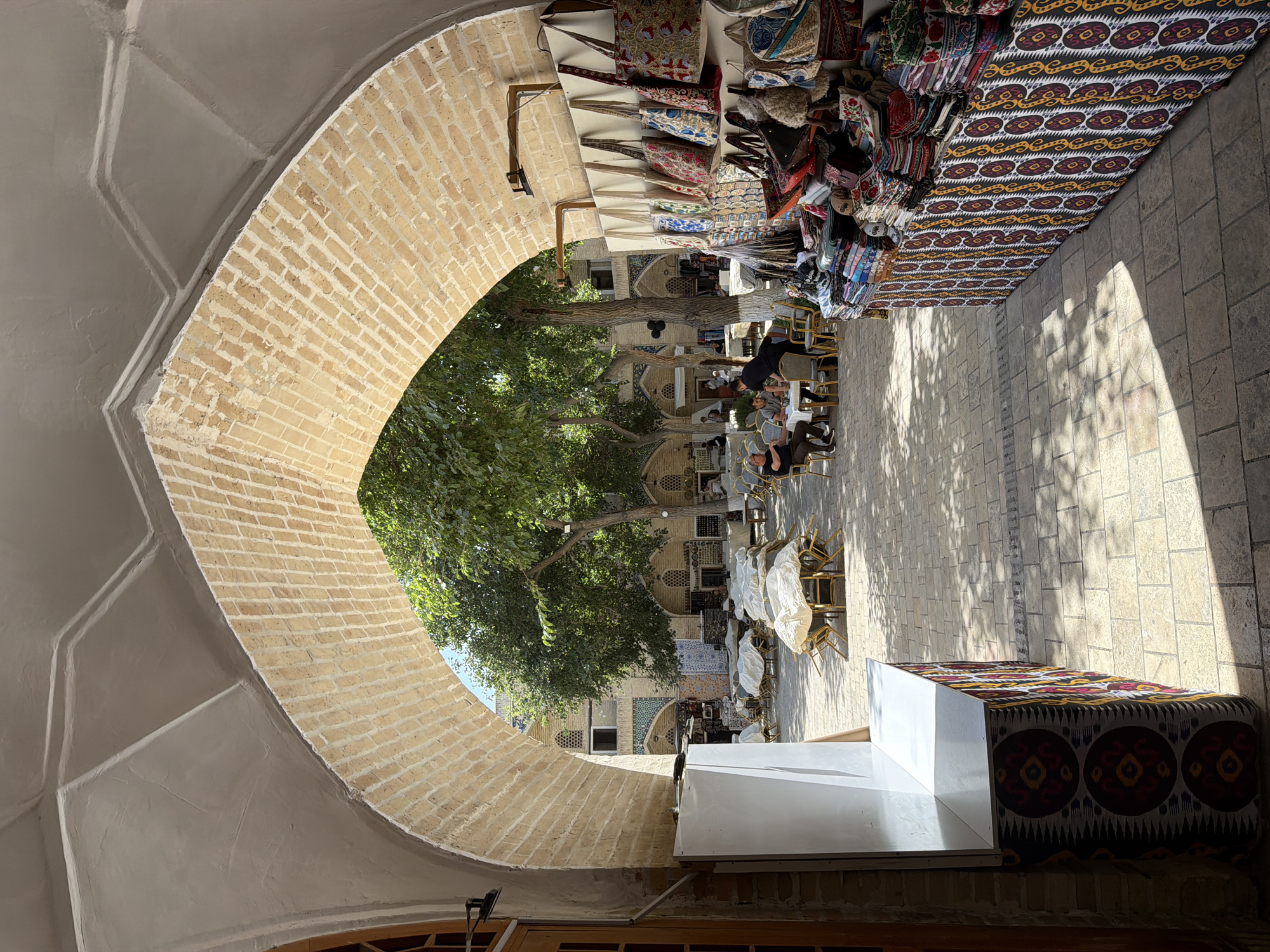 The image depicts a sunlit courtyard scene in Bukhara, Uzbekistan, observed from beneath a large, light-colored brick archway.
**Archway:** The archway's inner surface is composed of yellowish-beige bricks arranged radially. Above the brickwork, the arch's ceiling is covered in smooth, light-gray stucco with subtle raised geometric patterns.
**Foreground:**
* **Left:** A tall, plain white, rectangular display or storage unit stands beside a partially visible wooden door frame. Below and extending right, the end of a counter is covered in a dark fabric patterned with repeating circular motifs in yellow and red, characteristic of ikat.
* **Right:** A market stall occupies the inner wall of the archway. Numerous embroidered and intricately patterned fabric bags hang from hooks on a light vertical surface. Below these, a long counter, identical to the one on the left in its dark ikat patterning, displays stacks of folded textiles, scarves, and smaller merchandise. A dark, wall-mounted light fixture is positioned above the hanging bags.
**Midground (Courtyard):** The courtyard floor is paved with light-toned rectangular stone or concrete pavers. Direct sunlight illuminates a significant portion of the ground, casting pronounced shadows from trees and structures.
* **Seating Areas:** Multiple tables with chairs are distributed across the courtyard. Several tables are draped with white protective coverings, while others are uncovered.
* **People:** Towards the left-center, three adult males are seated around a square table, appearing to be conversing. One man wears a dark polo shirt, another a light collared shirt, and the third a dark vest over a light shirt. A male server in a dark uniform stands facing away from the camera, near their table. Additional indistinct figures are present further in the background.
* **Vegetation:** Several mature trees with full green foliage are interspersed within the courtyard, providing areas of shade.
**Background:** Traditional Central Asian buildings frame the far side of the courtyard under a clear blue sky. These structures feature light-colored facades, arched window openings, and subtle decorative elements, including patterned tilework visible on a structure to the left. Some buildings exhibit multiple stories.