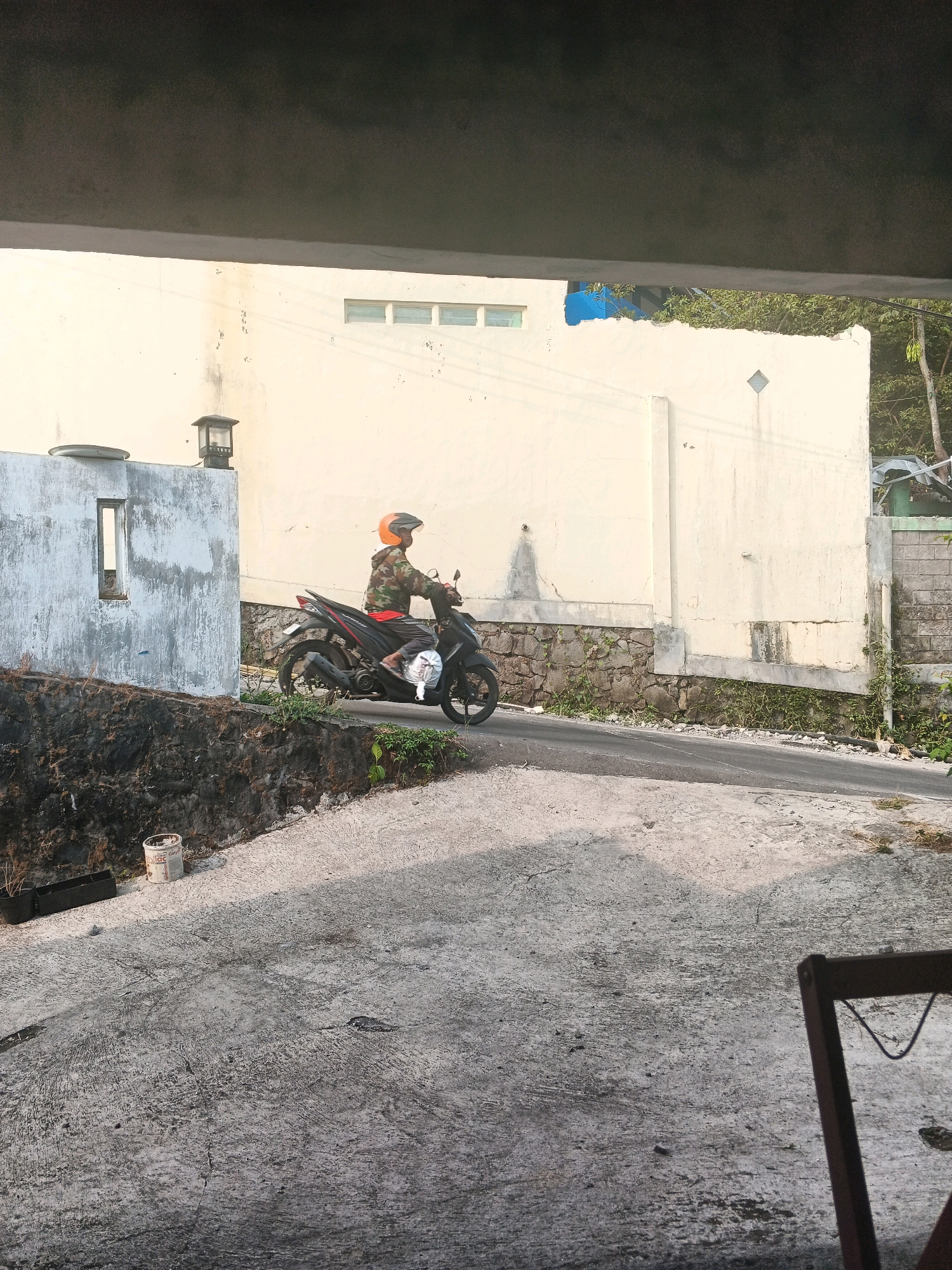 A person rides a motorcycle on a paved road in a village setting. The person is wearing a camouflage jacket and an orange helmet. The motorcycle is black with red accents. In the background, there is a light-colored building with a stone foundation. There is also a concrete structure with a small window and a lamppost. The road is bordered by a stone wall and vegetation. The ground in the foreground is also paved with concrete. The image appears to have been taken during the daytime.