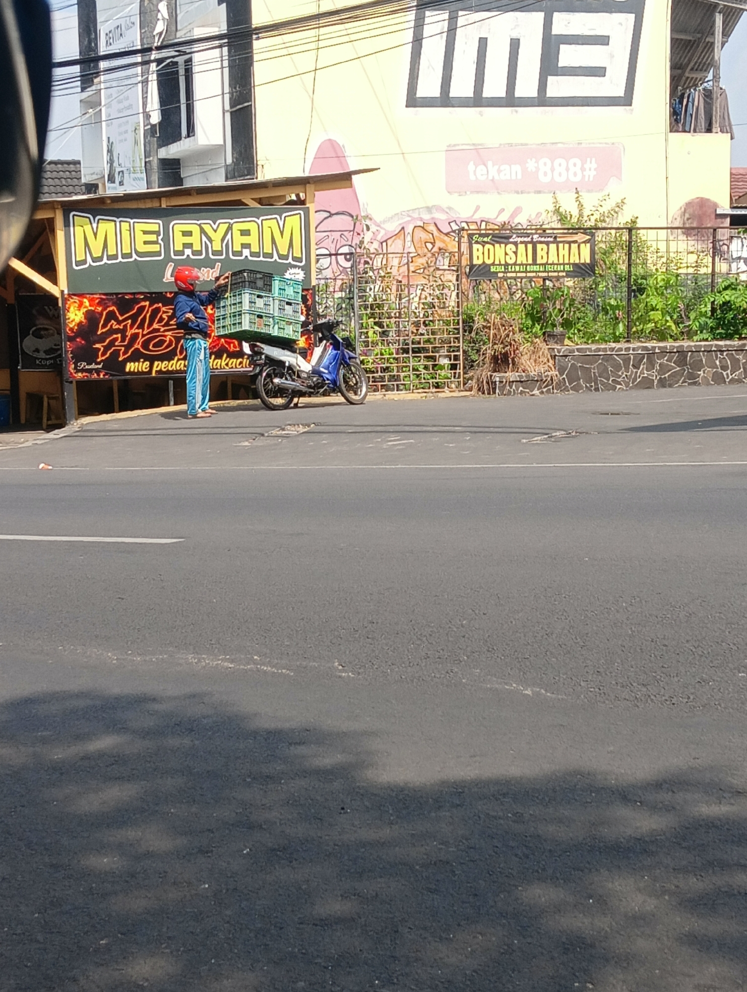 In Cijoho, Indonesia, a person wearing a red helmet and blue pants is loading green crates onto a blue motorcycle parked on the side of a road. The motorcycle has a metal rack on the back, holding several stacks of crates. A banner with the words "MIE AYAM" in large yellow letters is visible behind the person and motorcycle, along with other advertisements and graffiti on the building in the background. The road in the foreground appears to be asphalt, with some cracks and debris. The environment suggests a commercial area, possibly a food stall or small business.