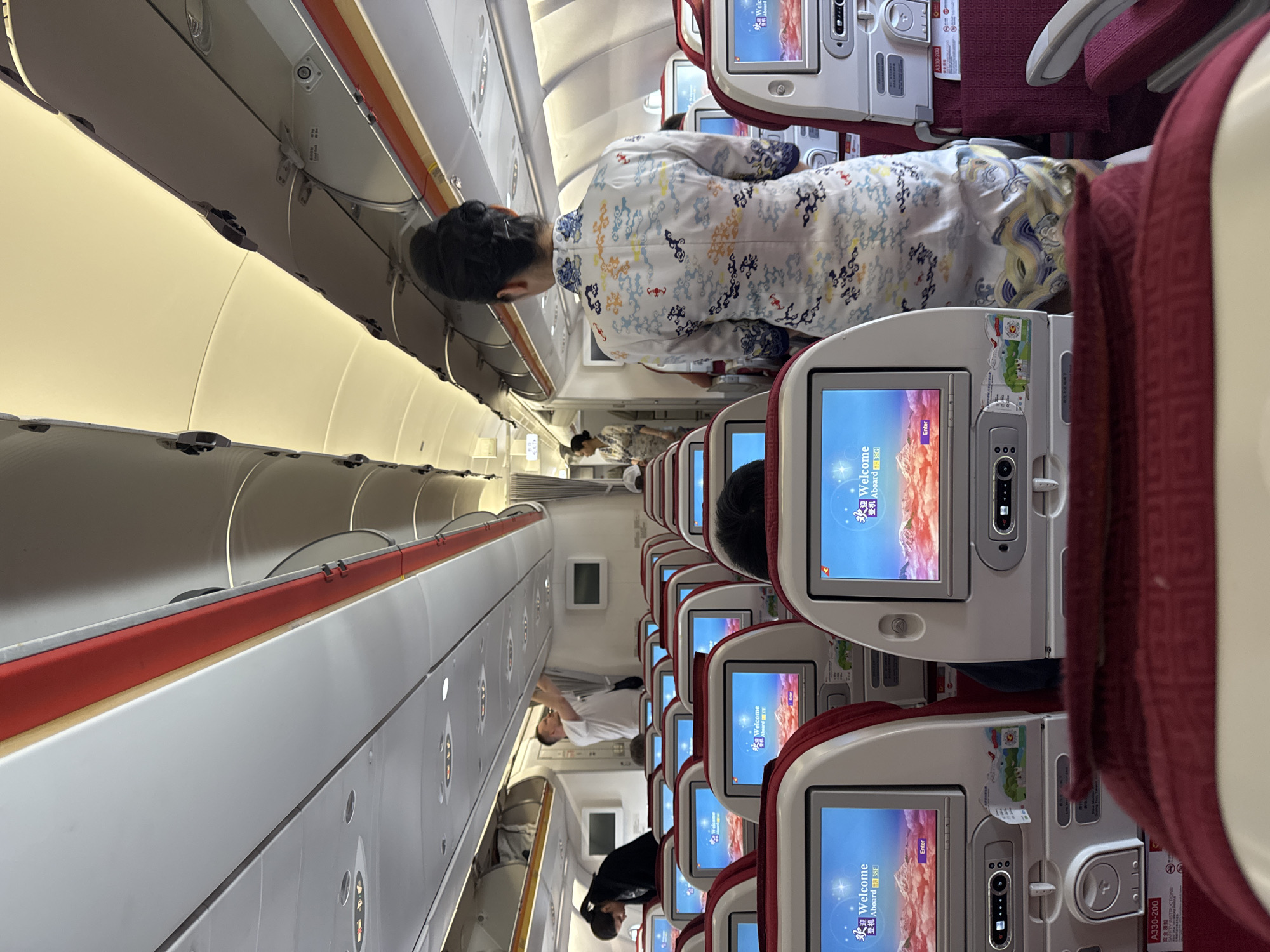 The image shows the interior of an airplane cabin, likely during boarding. The camera angle is from the perspective of a passenger looking forward down the aisle. Rows of seats are visible, each equipped with an entertainment screen displaying a "Welcome Aboard" message in both English and Chinese. Passengers are standing in the aisle, and flight attendants can be seen assisting them. The lighting inside the cabin is bright. Overhead bins are visible along the ceiling.