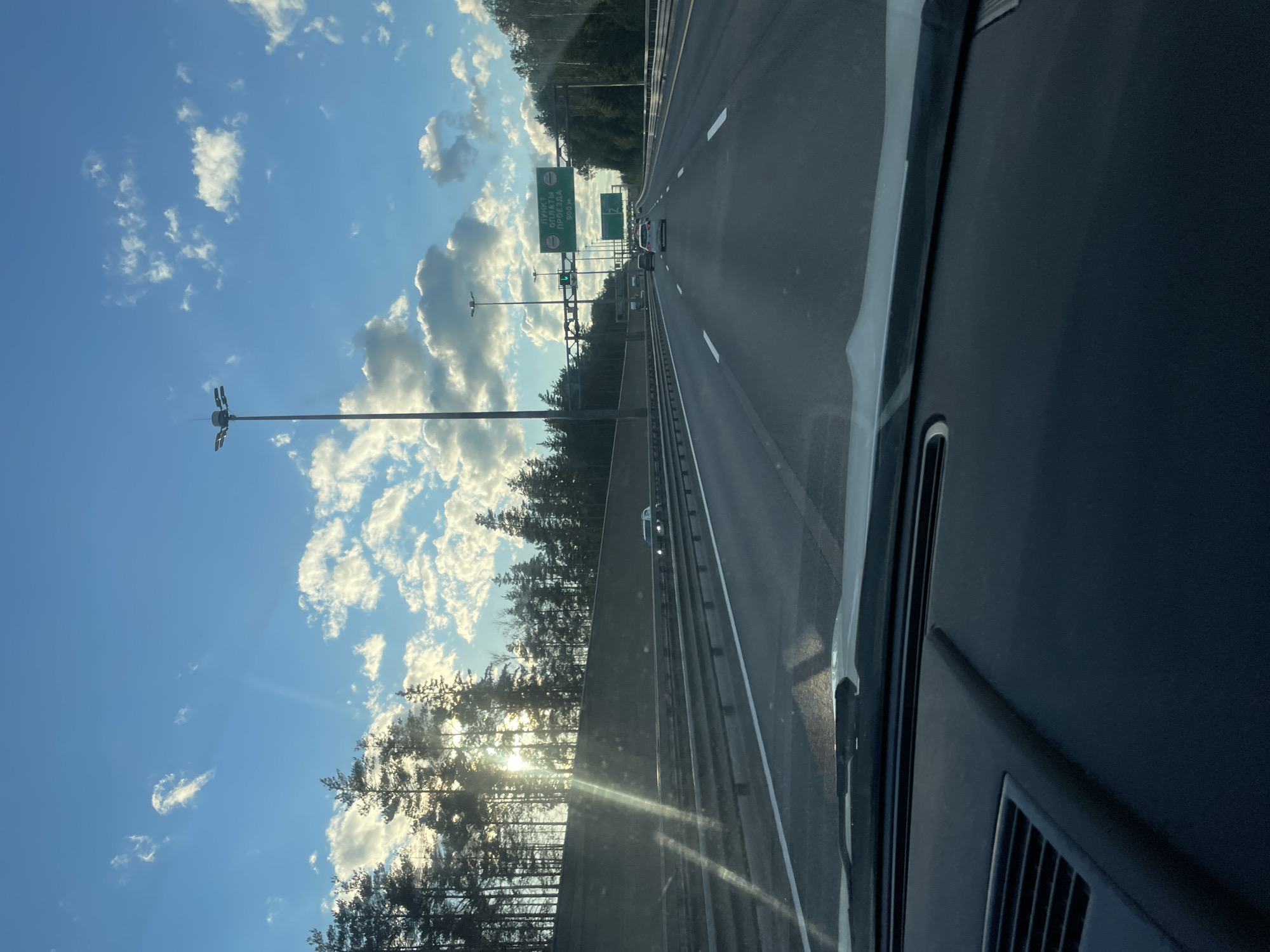 The image captures a perspective from inside a vehicle traveling on a multi-lane highway. The highway stretches into the distance, bordered by a concrete barrier on the left and a tree line on the right. Tall light poles are visible along the roadside. Several cars are driving on the highway in the same direction as the viewer. Overhead signs are present, one of which indicates "ПУНКТ ОПЛАТЫ ПРОЕЗДА" which translates to "Toll Payment Point" and a distance of "900 M". The sky is bright blue with scattered white clouds, and the sun is shining, casting rays through the trees and creating lens flare on the windshield. The foreground shows the dashboard and windshield of the vehicle. The scene appears to be set in Pesochny, Russia, given the Cyrillic text on the signage.