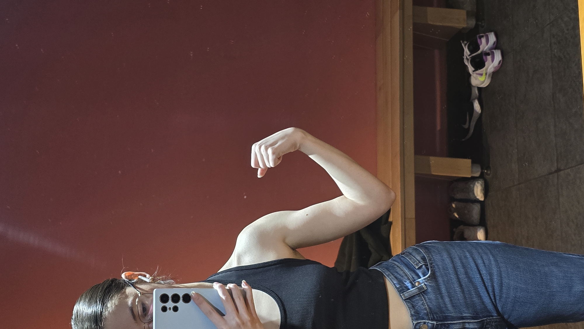 A woman is taking a selfie in a mirror. She is flexing her right bicep. She is wearing a black tank top and blue jeans. She is holding a smartphone in her left hand. The background includes a red wall, a wooden frame, a shelf with shoes, and a dark tiled floor. The image was taken inside a building. It is impossible to determine if this image was taken in Stara Zagora, Bulgaria without additional information.