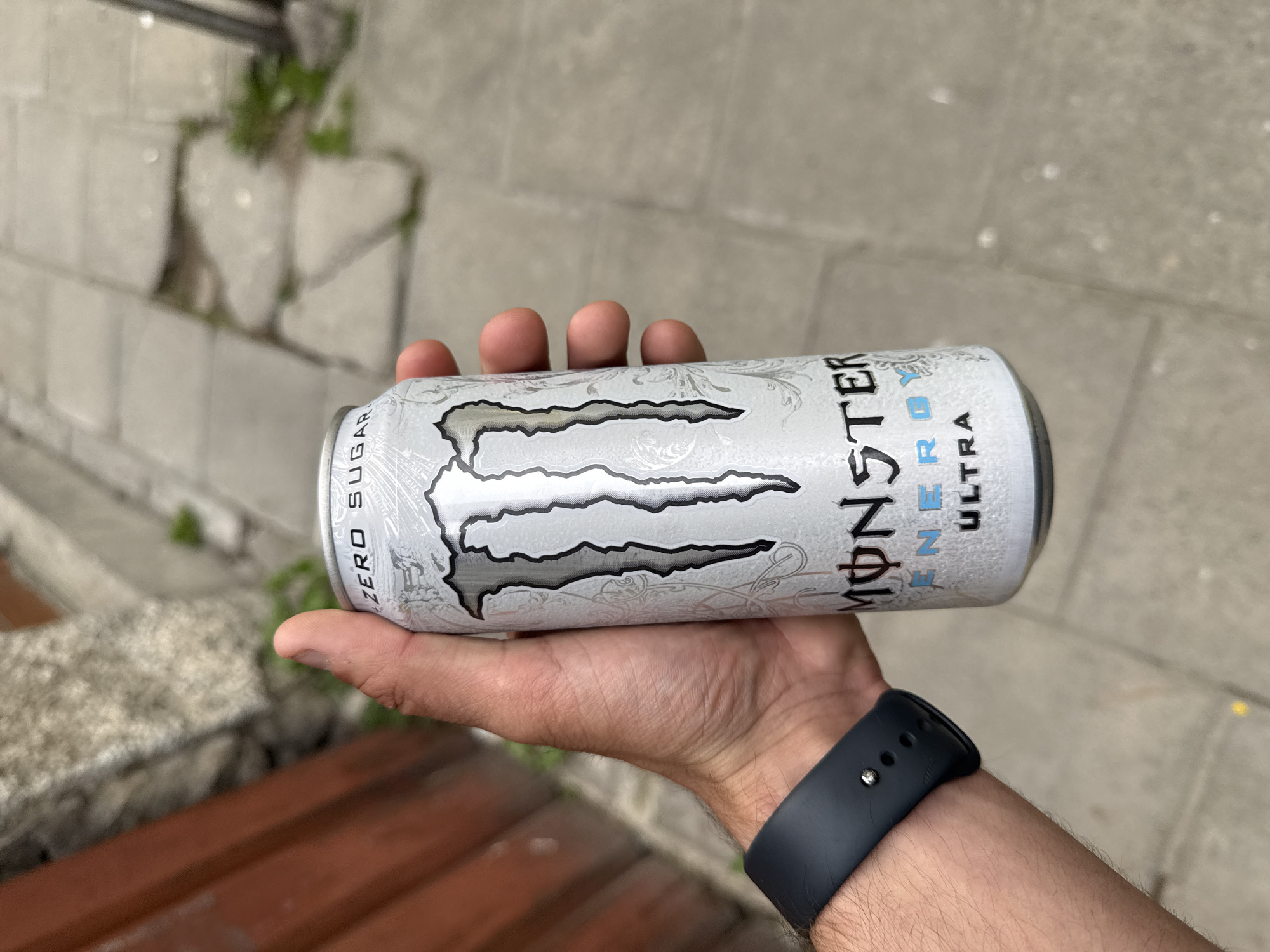 A medium-shot image depicts a fair-skinned male holding a white Monster Energy Ultra Zero Sugar can in his left hand. He is wearing a black smartwatch. The background consists of a stone wall and a partially visible wooden bench. The image appears to be taken outdoors in a public space. The specific location in Shumen, Bulgaria is not evident from the visual information provided.