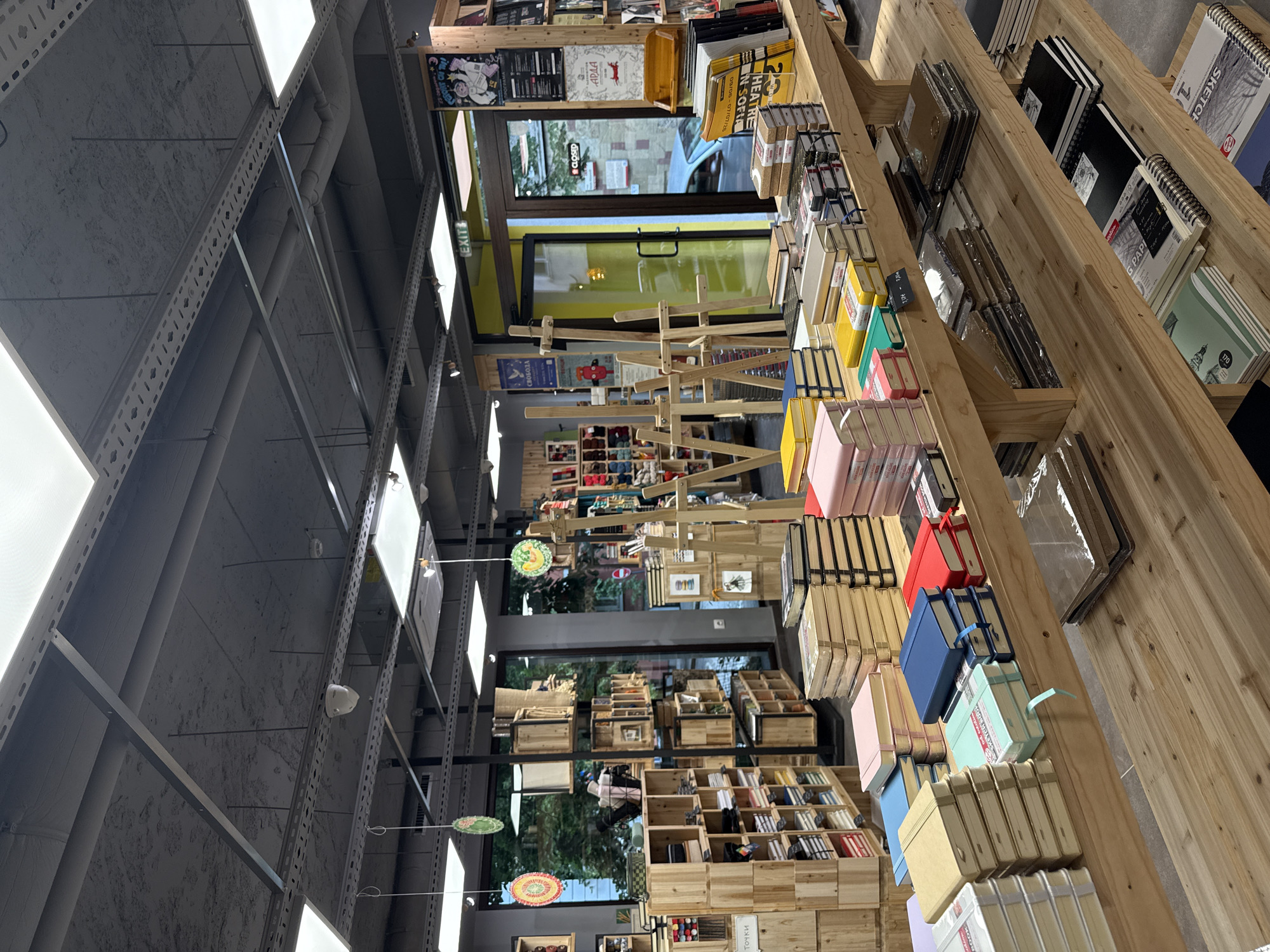 The image displays the interior of an art supply or craft store, characterized by a modern, industrial design. The perspective looks along rows of wooden shelving units that are packed with merchandise. The ceiling features exposed concrete with visible pipes and linear LED panel lighting, casting bright light on the store.
The shelves are predominantly made of natural wood and hold a vast assortment of products. These include numerous sketchbooks, notebooks, and binders in various sizes and colors, as well as loose paper pads and art sets. Crafting supplies, such as yarns, are also visible in wooden crates and cubbies. Several wooden easels are interspersed among the products, possibly for display.
Towards the middle and back of the store, a large glass door or window reveals reflections of the outside, including a parked car and building exteriors. A "CLOSED" sign is visible on this door, suggesting the store is not currently open for business. An "EXIT" sign is also present nearby. Posters adorn the walls, one featuring "HEATRE N SOFIA" with dates "08/06-07/07/28," another showing "АРДА" (Arda), and a third with "СВОБОДА" (Svoboda - Freedom). Round, colorful decorations hang from the ceiling at various points.
Other visible text on products includes "SKETCH," "G PAPI," "DIAD," and numbers like "170." A label on some crafting supplies reads "Точки" (Tochki - Dots/Points). The scene captures the diverse inventory and clean, organized layout of a specialty store in Sofia, Bulgaria, as indicated by the "Sofia" reference on a poster.