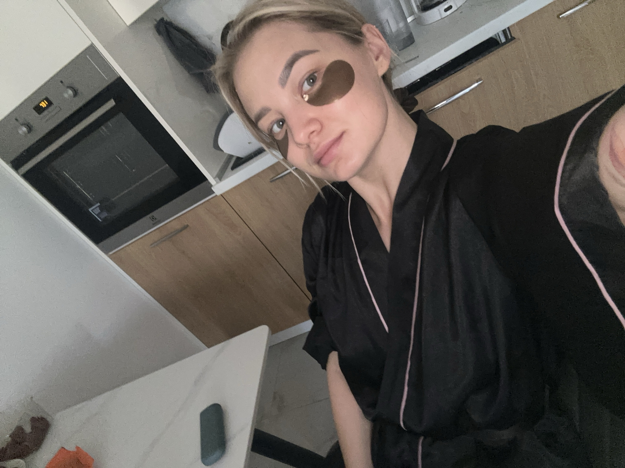 A woman with light hair and eye masks is taking a selfie in what appears to be a kitchen. The kitchen has light-colored cabinets, a grey oven, and a white countertop. She is wearing a black robe with pink piping. Based on the style of the kitchen and general appearance, this could be a residential setting. There is no conclusive information in the image to place the location in the city of Budva, Montenegro.