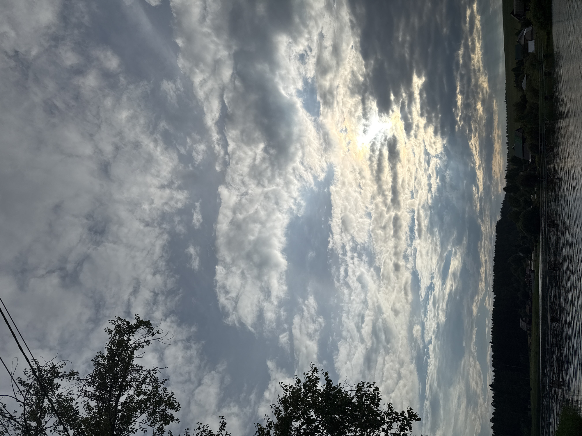 The image captures a serene landscape scene, likely near the village of Luzhkovo, Russia, with a focus on the sky and a body of water. The sky is filled with a dramatic arrangement of clouds, with patches of sunlight breaking through, creating a textured and dynamic atmosphere. The lower portion of the image reveals a dark, tranquil body of water, its surface rippled by a gentle breeze. Beyond the water, a dense forest of dark green trees forms a natural backdrop. Scattered among the trees are a few small buildings or houses, suggesting a rural settlement. A few branches and leaves from a tree frame the upper left corner of the image. The overall impression is one of natural beauty and peacefulness.