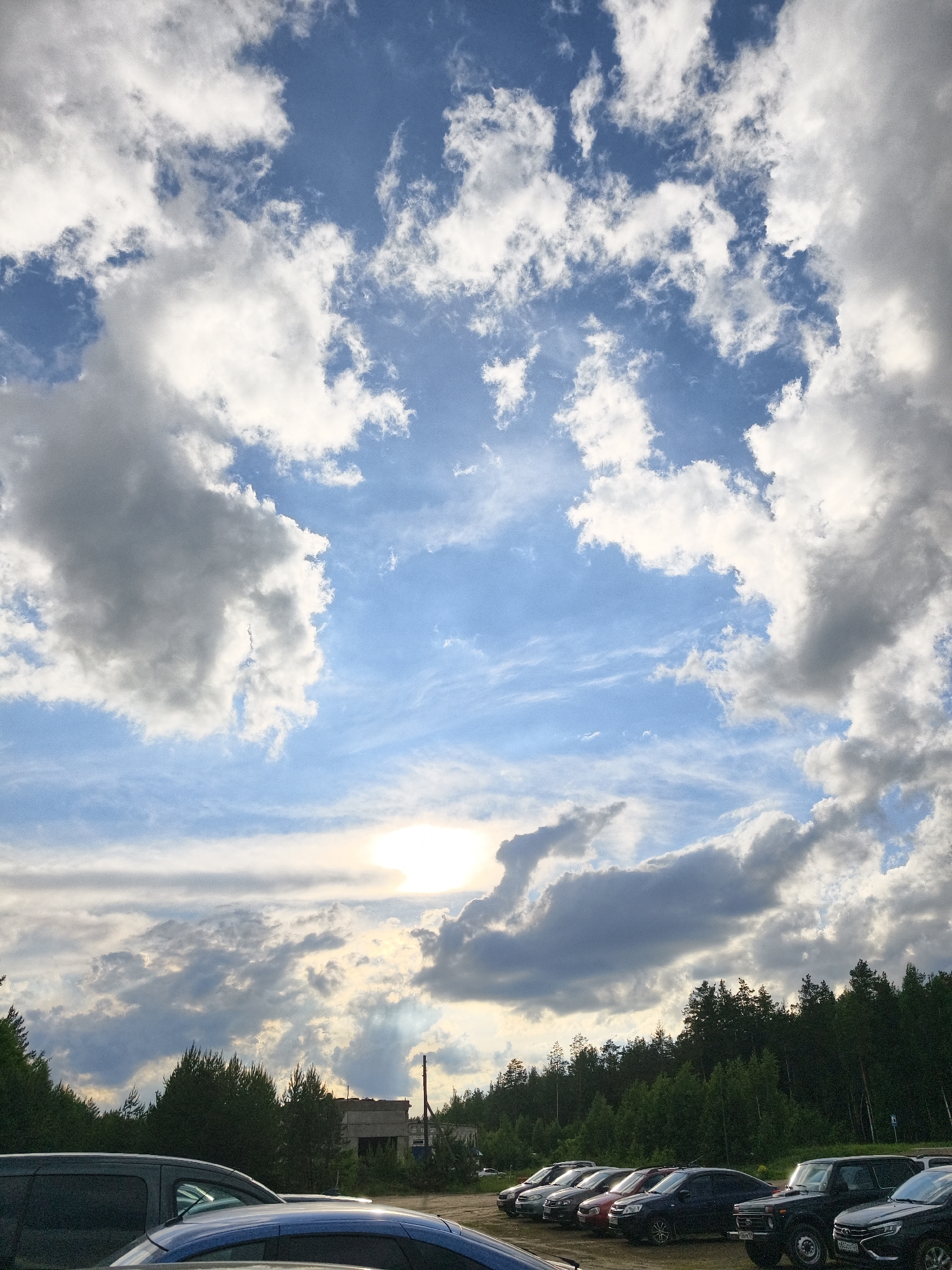 The image shows a parking lot in Noviy, Russia. Several cars of various makes and models are parked in a row, including a dark SUV, a red sedan, and several other vehicles. Behind the cars is a line of trees, and behind the trees is a building. The sky is blue with a mix of white and gray clouds. The sun is partially obscured by the clouds, casting a bright glow in the lower center of the image. The lighting suggests it is either morning or late afternoon.