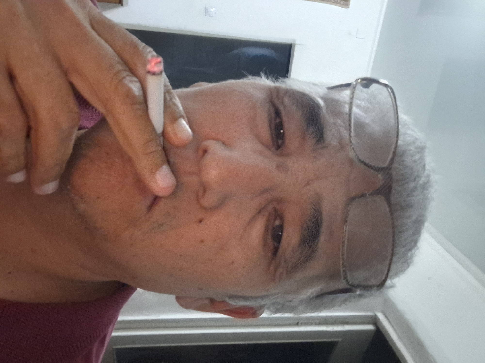 A close-up, self-taken photograph shows a man with graying hair and glasses holding a lit cigarette to his lips. He is wearing a maroon polo shirt. The background appears to be an indoor setting with a white wall and a dark doorway or window. The lighting is dim. There is no discernible information in the image to indicate a specific location in the city of Almaty, Kazakhstan.