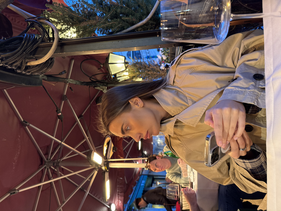 The image depicts an evening or nighttime scene in a semi-enclosed outdoor dining area of a restaurant in Venice, Italy.
**People:**
In the foreground, a young adult female with long, straight brown hair is seated at a table, shown in profile facing right. She wears a tan trench coat over a plaid shirt and holds a silver-colored smartphone with a clear case in both hands; a ring is visible on her left ring finger. To her immediate left, a portion of another individual's arm and a wine glass filled with red wine are visible on the same table. Further into the background, a second female with light-colored hair, wearing a light green top, is seated at another table. A green bottle stands on her table. A male figure in a dark jacket is partially visible in the distant left background.
**Objects and Environment:**
The dining area is sheltered by a large, dark red patio umbrella structure, from which two active overhead heaters emit a warm glow. Tables are covered with white tablecloths and set with glassware. A prominent dark metal pole stands on the right, adorned with coiled black electrical cables and an ornate, cream-colored lantern emitting a warm light. An evergreen tree or bush is situated to the far right against a dark blue sky, with a second, smaller lantern partially visible behind it. An empty, clear wine glass with a thin stem is positioned on the white tablecloth in the foreground to the right of the primary subject. In the dimly lit background, architectural elements resembling arched windows and columns of a large classical building are faintly discernible, consistent with Venetian cityscape.
**Actions and Interactions:**
The primary subject is holding her smartphone and looking towards the right side of the frame. Other visible individuals are seated at tables, consistent with dining. No direct interactions between the individuals are captured.