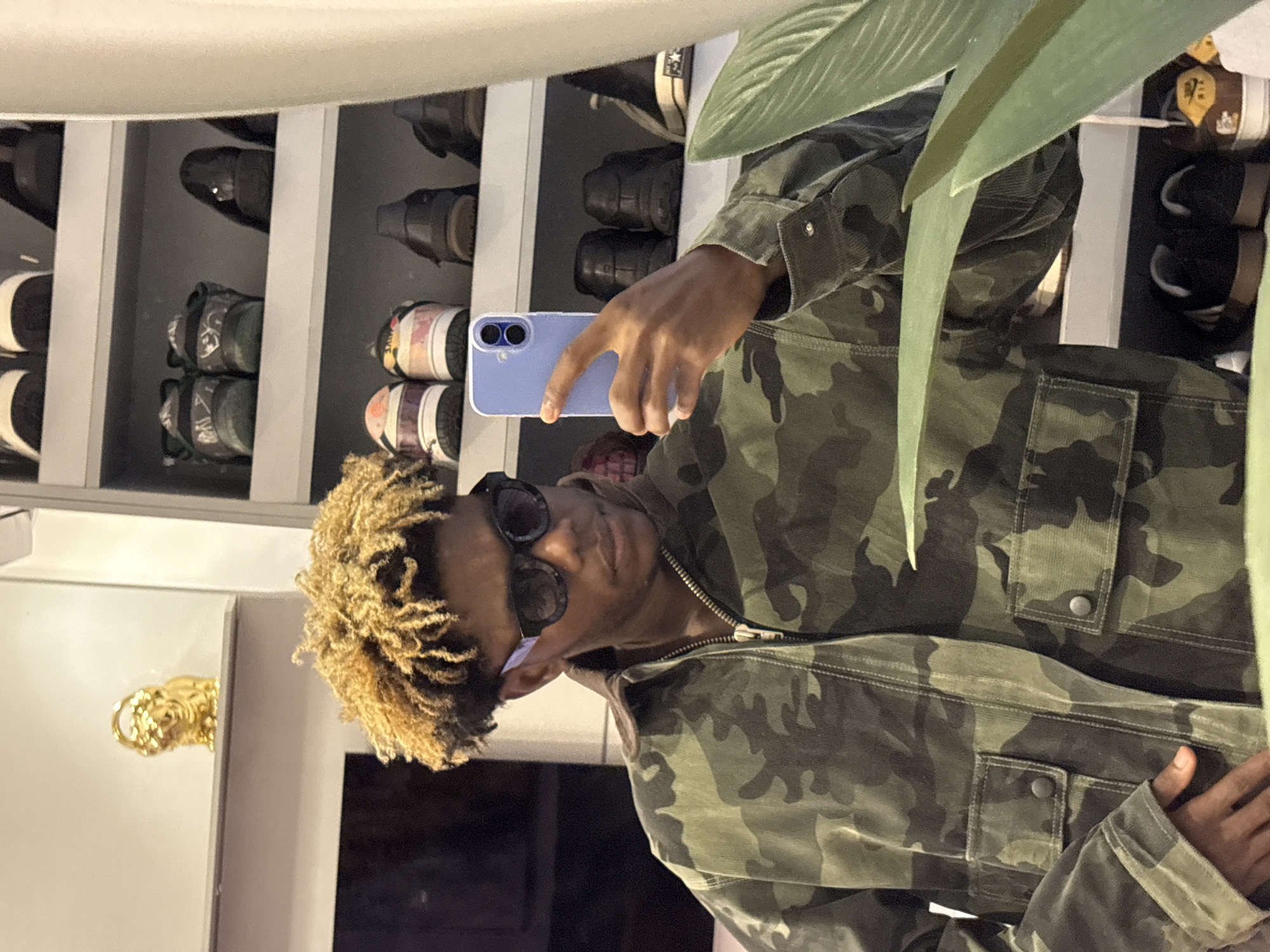 A young Black male with textured, light brown hair and dark sunglasses is depicted taking a mirror selfie indoors. He wears a camouflage-patterned jacket with a zippered front and button-flap chest pockets. His left hand holds a light blue smartphone, pointed towards the unseen mirror. The background on the left features a white wall with a shelf holding a small golden figurine resembling a lion. The background on the right consists of a shelving unit displaying multiple pairs of shoes, including dark dress shoes, sneakers with various designs (e.g., green/black, patterned, brown/tan), and a pair of black Converse All-Stars with a white star logo visible. A large green leaf from an indoor plant partially obstructs the lower right foreground. The specific location within the city of Igboefon, Nigeria, cannot be determined from the visual information provided.