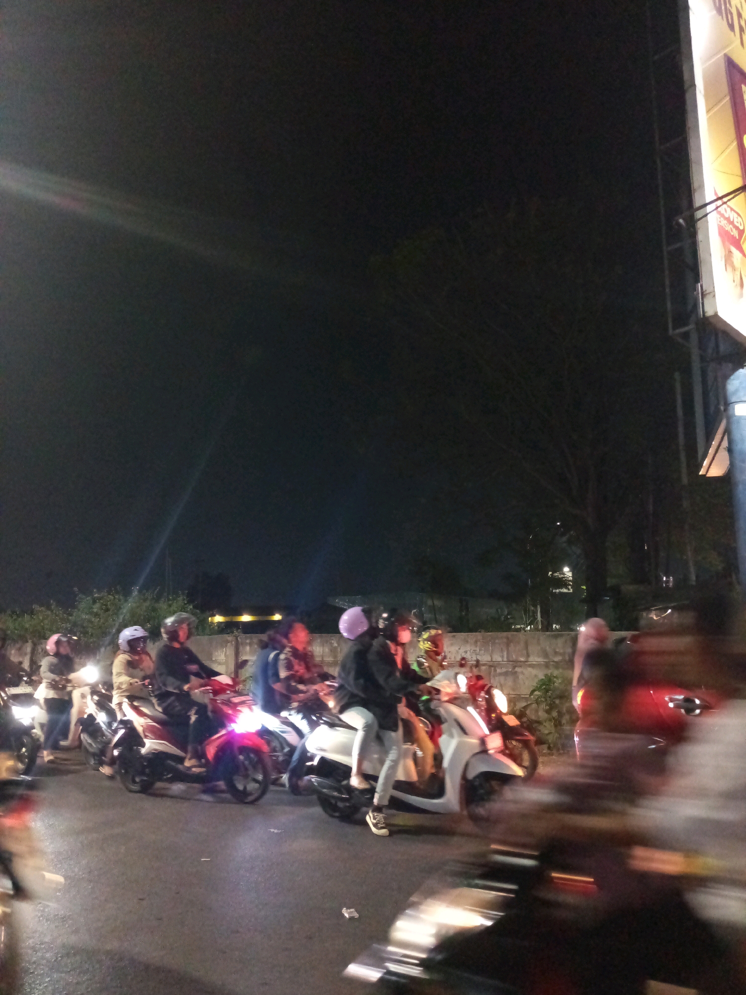 In Bekasi, Indonesia, a group of people on motorcycles are captured in a nighttime street scene. The image is blurry, suggesting motion, and is illuminated by streetlights and the headlights of the vehicles. The people are wearing helmets, and some are wearing jackets and casual clothing. The motorcycles are a mix of modern scooters. The background shows a dark sky, trees, and a building with a lit sign, possibly a store or advertisement. The road appears to be wet, indicating recent rain or damp conditions. The overall atmosphere is urban and active, with people on their way somewhere in the night.