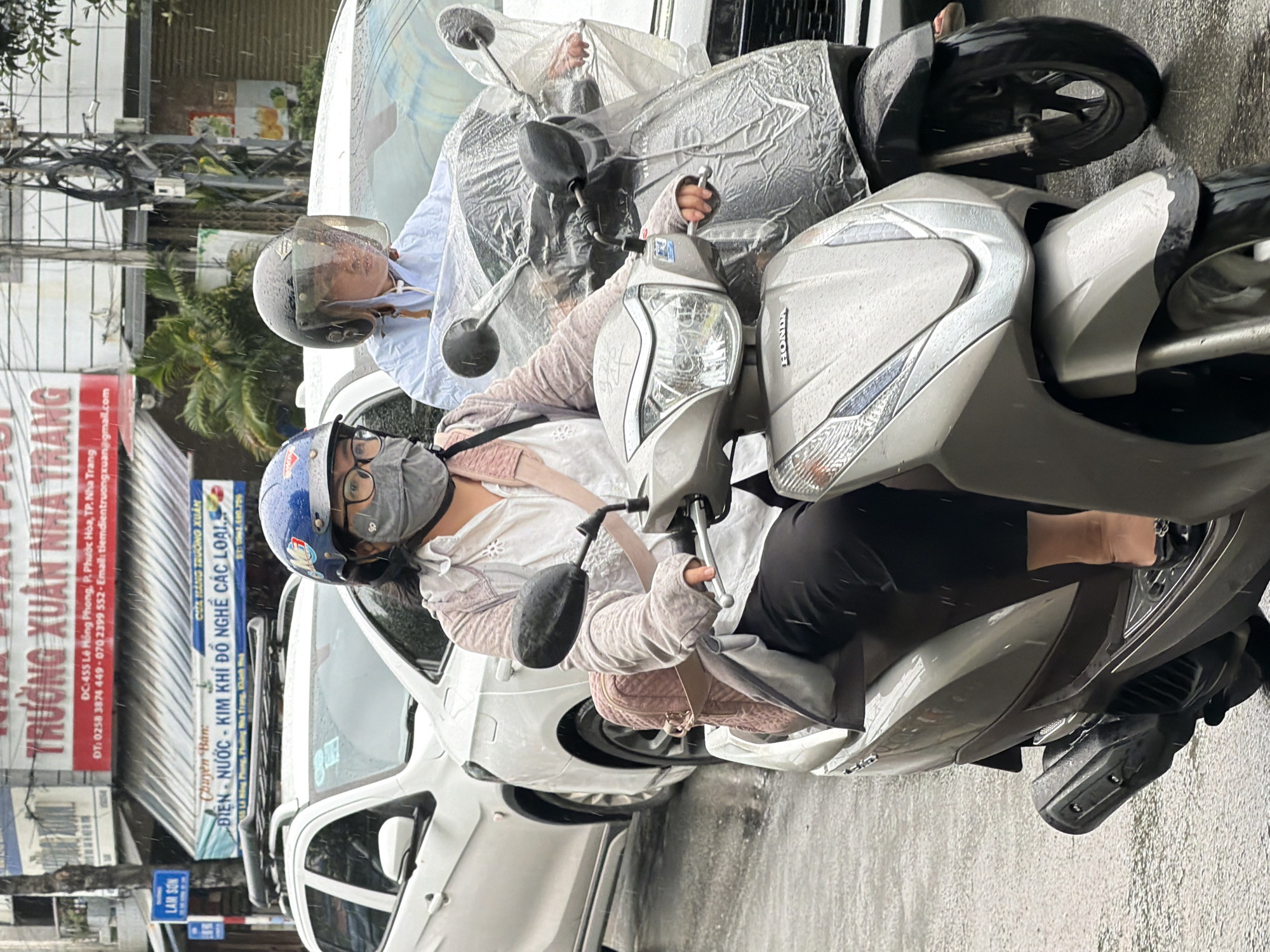 The image captures a street scene in Nha Trang, Vietnam, likely on a rainy day, indicated by the wet asphalt and raindrops visible. In the foreground, two individuals are riding scooters. The scooter on the left is ridden by a person wearing a blue helmet with a transparent face shield, glasses, and a grey face mask. They are also wearing a light-colored long-sleeved shirt and dark pants, and a quilted pink backpack is visible on their back. The scooter itself is silver and appears to be a Honda. The scooter on the right is mostly covered by a clear plastic sheet, with a person wearing a white shirt and a dark helmet visible underneath. The background shows buildings with signs, including one in red with Vietnamese text that appears to read "TRUONG XUAN NHA TRANG," suggesting a business or establishment. Another sign below it has more text and contact information. White cars are parked or moving in the background. The overall scene depicts daily life in a Vietnamese city during inclement weather.