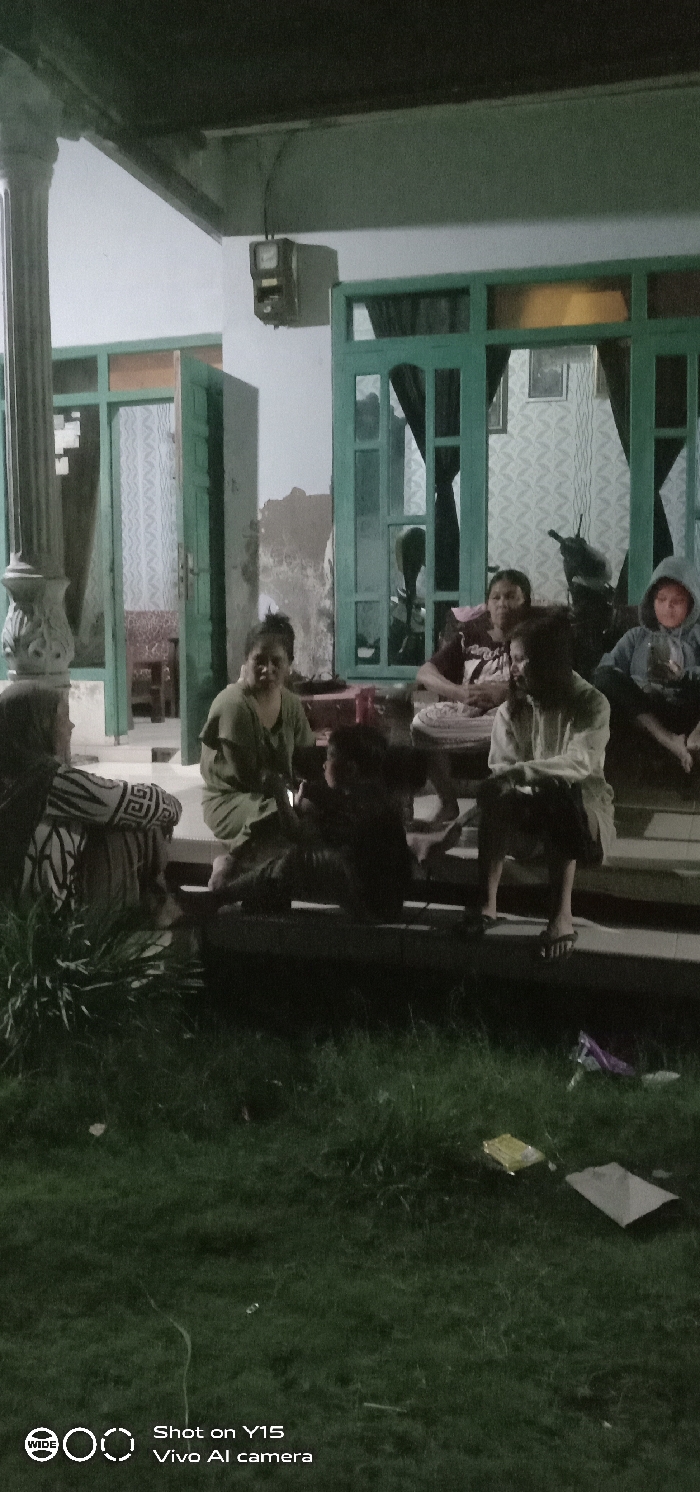 A group of people is gathered on the porch of a house at night. The house has green trim and large pillars. At least six individuals are visible, seated on the steps or nearby. The scene is dimly lit, with shadows obscuring some details. In the background, there's a glimpse of a room through an open door and window. The grass in front of the porch is littered with trash.