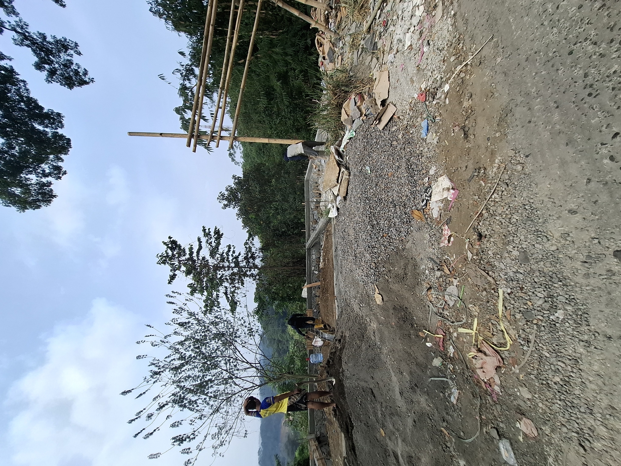 The image depicts a construction site in Cibadak, Indonesia, under a cloudy sky. Three individuals are engaged in construction work. One person, wearing a yellow and blue shirt and black shorts, stands near a pile of dirt, holding a shovel. Another person, dressed in dark clothing and yellow boots, is working with cement in a foundation area. The third individual, in dark clothing, is also involved in construction activities. The site is filled with construction materials, including gravel, cement, bamboo scaffolding, and various debris. The background shows lush green vegetation and a mountainous landscape, typical of Cibadak's surroundings. The ground is a mix of dirt and gravel, with some scattered refuse.
