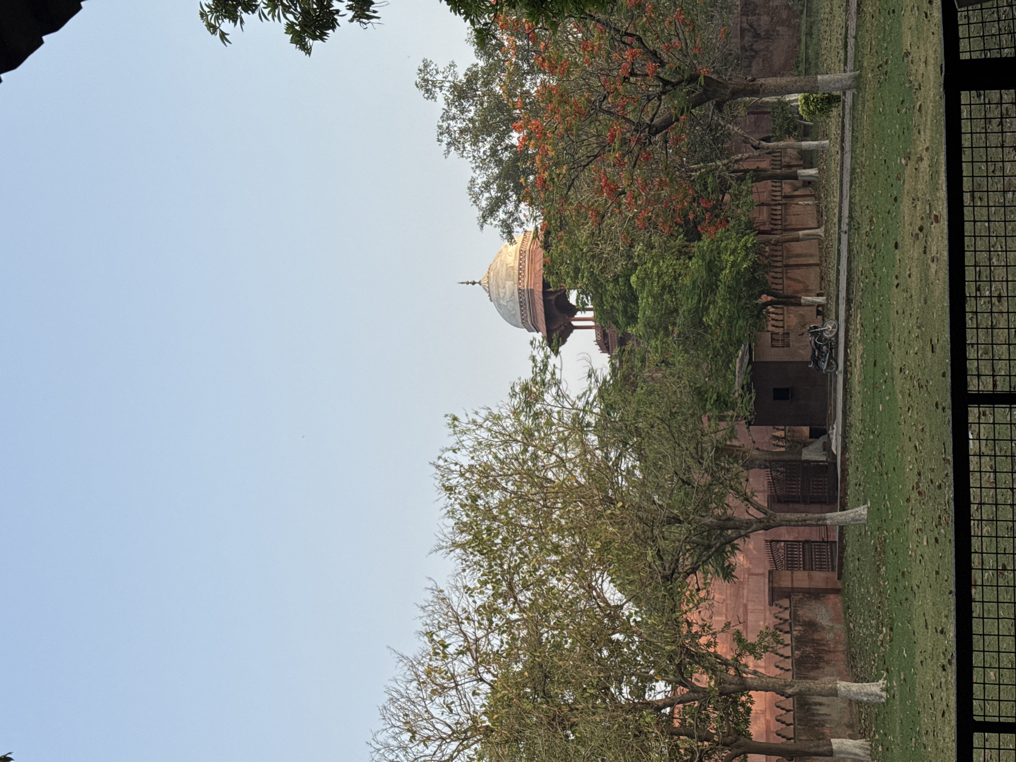 The image presents a view of an architectural structure with a prominent dome, likely located in Agra, India. The building features red sandstone elements, and the dome is painted white, with golden accents at its peak. The structure is partially obscured by trees with varying degrees of foliage, some with red flowers. A well-maintained green lawn stretches from the base of the structure toward a wire fence, which appears to mark the boundary of the site. A parked motorcycle is visible near the base of the building, suggesting possible accessibility. The sky is clear.