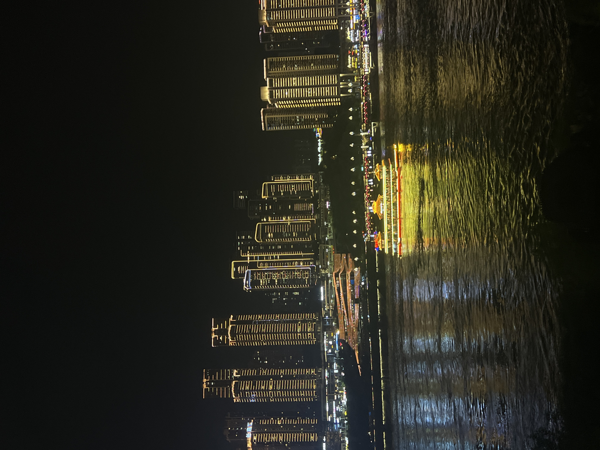 This is a nighttime scene at Jiangbei City Street, China, featuring a cityscape with illuminated skyscrapers and a river in the foreground. A large tour boat with glowing yellow and red lights is moving on the river, casting reflections on the water. The buildings are lit with golden and white lights, creating a vibrant urban display. The river is dark, with ripples reflecting the city lights. The overall atmosphere is one of a bustling, illuminated city at night.
