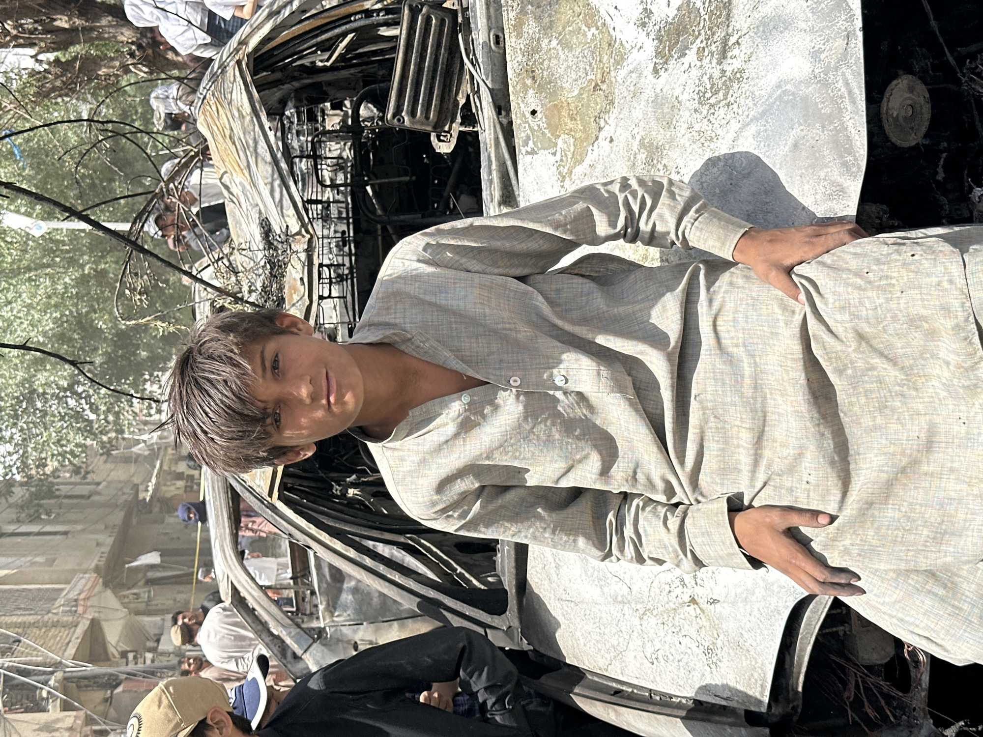 A young person stands near a severely damaged car, potentially in Quetta City Tehsil, Pakistan. The car's charred engine and frame are visible, indicating fire damage. The background includes a partially destroyed building and several people observing the scene. The young person is wearing traditional clothing and appears to be looking at the camera. The overall scene suggests a possible explosion or attack.