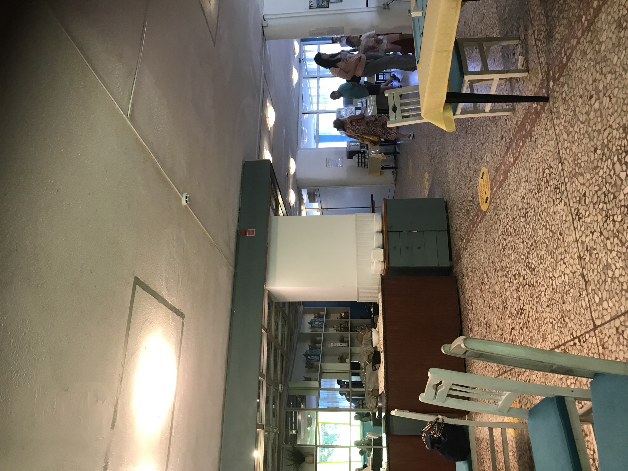 The image depicts the interior of a self-service restaurant or canteen in Varna, Bulgaria. The environment is characterized by a spacious, well-lit dining area with a textured, mosaic-like floor and a suspended ceiling with recessed lighting. Several tables and chairs are arranged throughout the space, some covered with yellow tablecloths. In the background, people are seen queuing for food or walking, suggesting an active service period. The decor includes shelving units stocked with decorative items and plants, reflecting a casual and functional setting. There is a buffet-style counter visible with various food serving stations. The overall impression is of a busy, casual dining establishment.