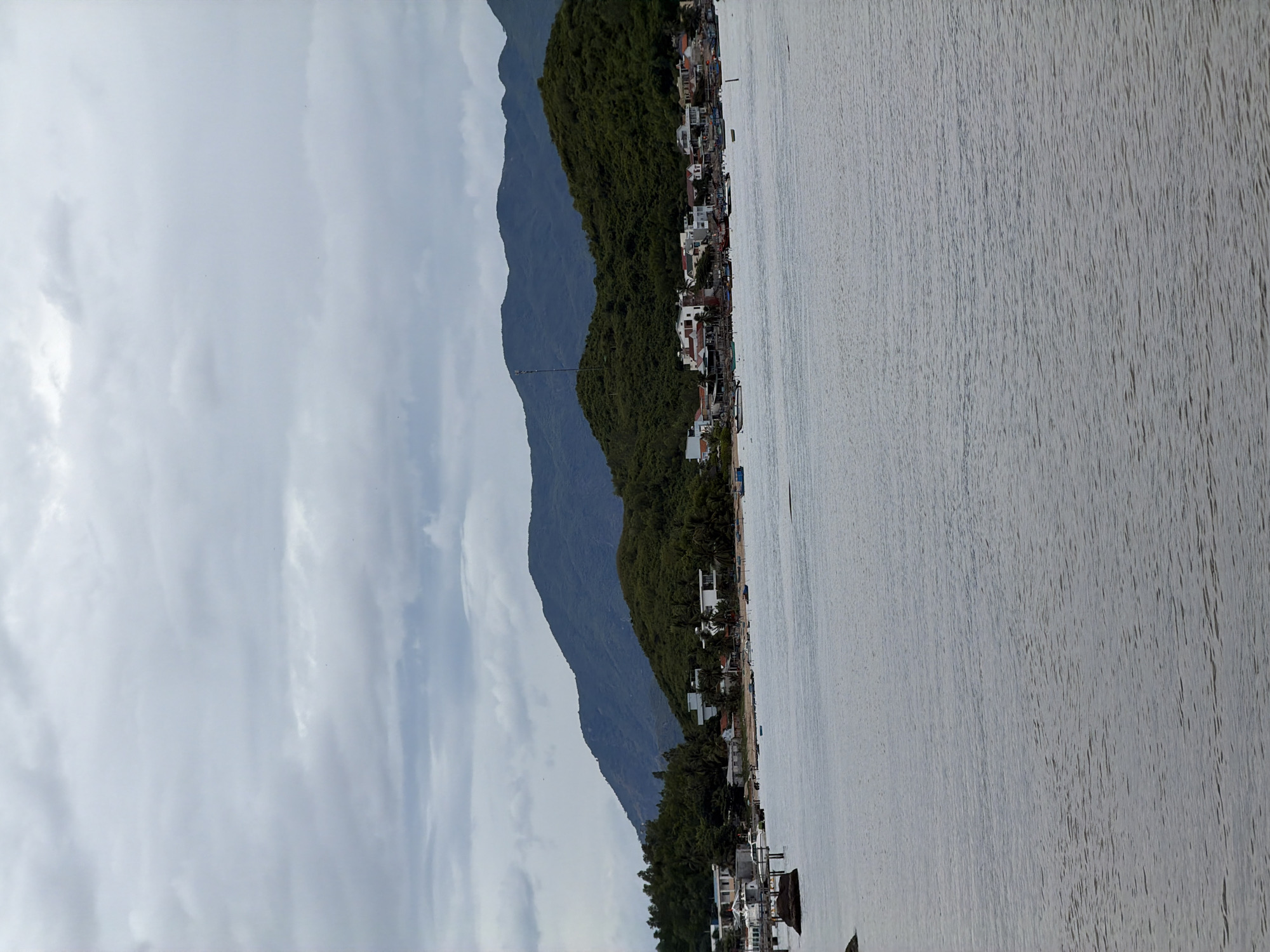 The image captures a serene coastal scene in Bắc Nha Trang, Vietnam. In the foreground, a body of calm, grey water stretches across the frame, its surface rippled by a gentle breeze. A few small boats are visible on the water, some anchored near the shore and others further out.
The middle ground showcases a picturesque coastline. Lush green hills, covered in dense vegetation and dotted with palm trees, rise majestically, forming a backdrop. Nestled along the shoreline is a small village, characterized by clusters of white and red-roofed buildings. The architecture appears to be a mix of modern and traditional styles, with some houses featuring balconies and awnings. A few larger buildings, possibly hotels or resorts, are also visible. Several more boats are moored along the beach, some of them appearing to be fishing vessels.
In the distance, the horizon is marked by a series of layered, dark blue mountains, their peaks softened by the hazy atmosphere. The sky above is overcast, with a thick layer of grey clouds that diffuse the light, creating a subdued and tranquil ambiance. There are no people clearly visible in the image, contributing to the overall sense of quietude. A communication tower stands tall on one of the green hills, a subtle sign of human infrastructure within the natural landscape.
