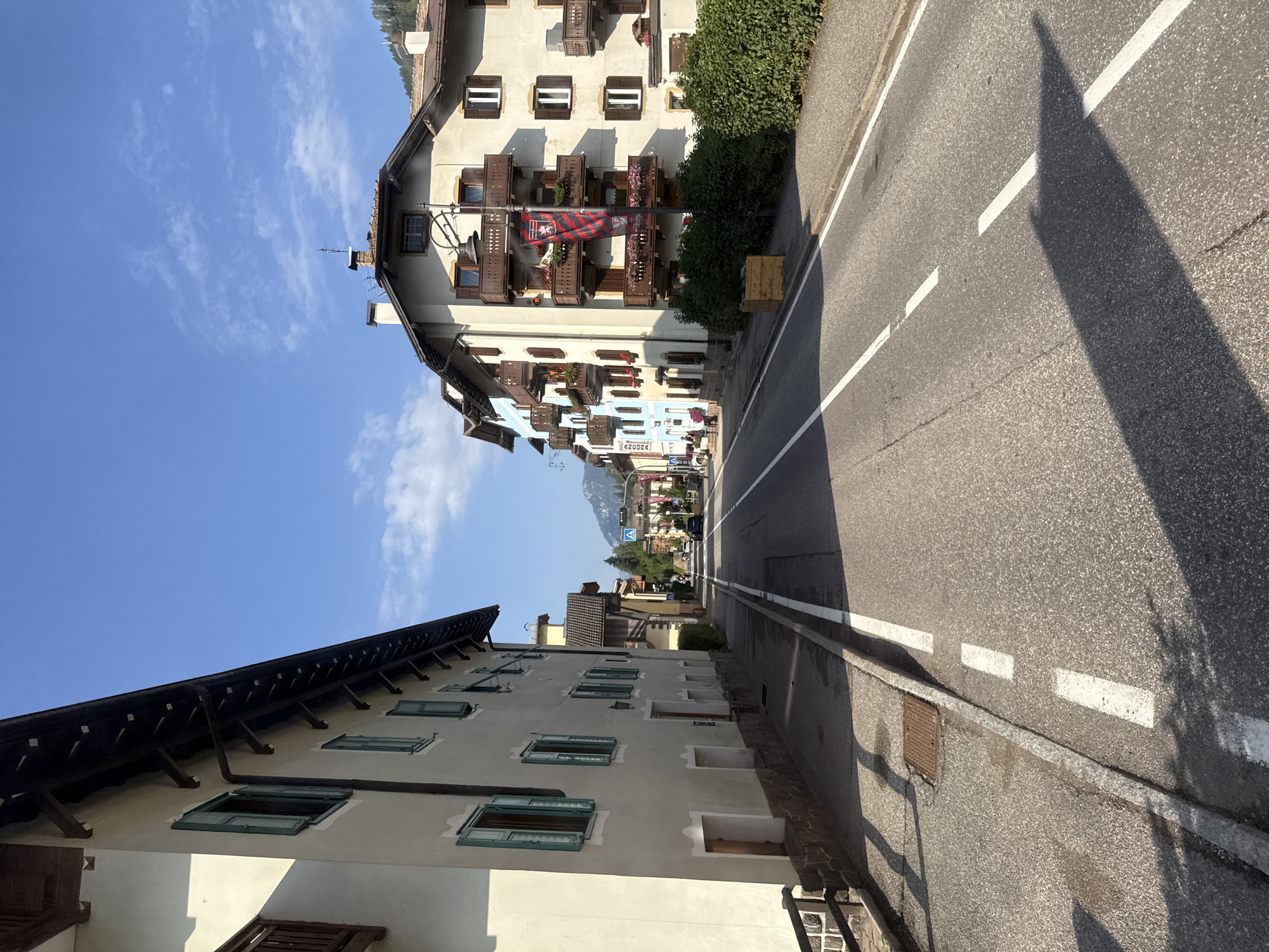 The image shows a street scene in Moena, Italy, on a sunny day with a clear blue sky and scattered white clouds. The street is paved and marked with white lane lines. On the left side of the street are the facades of buildings with light-colored walls, wooden balconies, and green shutters. A dark wooden fence runs along the sidewalk. On the right side of the street, buildings with similar architectural features are visible, including balconies with flower boxes and a banner hanging from one building. There are also several green bushes lining the sidewalk. In the distance, the street curves and appears to lead towards a mountainous landscape. A few vehicles are present on the road. The overall impression is of a well-maintained street in a European town.