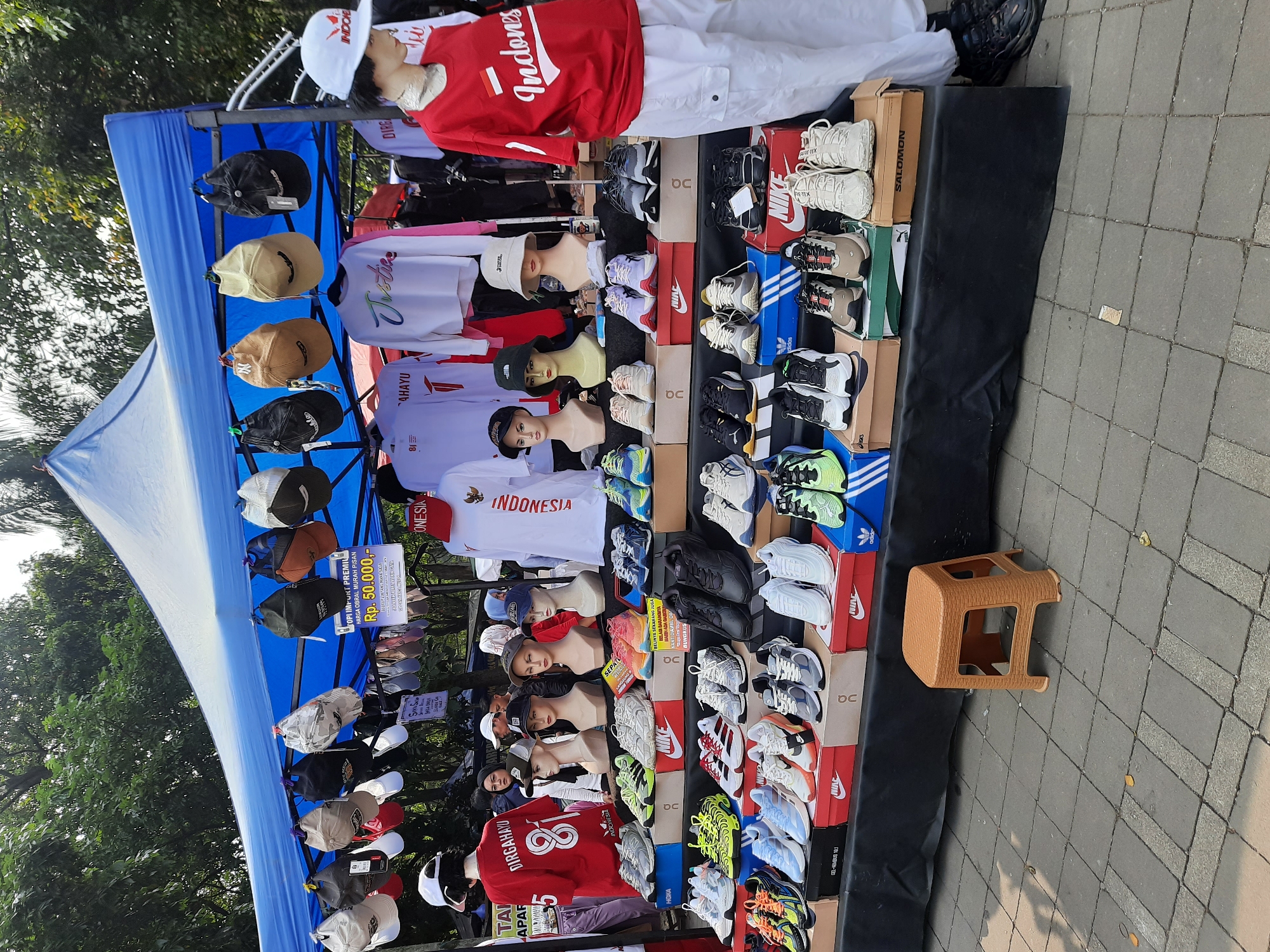 This image depicts an outdoor stall selling clothing and footwear, likely located in a public area of Bandung City, Indonesia. A blue tent provides shade over a display table filled with numerous pairs of sneakers, predominantly athletic shoes in various colors and styles. The sneakers are arranged on shelves and in shoe boxes, with brands like Nike and Adidas visible. Above the footwear, a collection of caps and bucket hats are hung from the tent's structure. Mannequins, dressed in t-shirts and displaying heads with hats, are positioned behind the shoe display. One mannequin on the right wears a red t-shirt with "Indonesia" printed on it in white lettering, along with white pants. The stall is set up on a paved ground, possibly a sidewalk or a market square. In the background, trees and foliage suggest a location with greenery. The overall scene is that of a street market or a temporary retail setup.