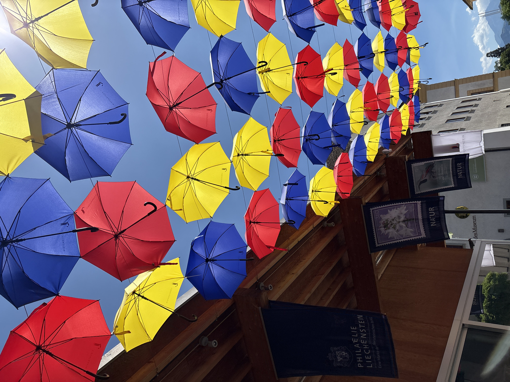 An outdoor shot captured at eye-level shows an art installation of red, yellow, and blue umbrellas suspended above a street. The umbrellas are arranged in rows, creating a vibrant canopy against a clear blue sky. Below the umbrellas, a wooden awning extends from a building, adorned with two dark blue banners. The left banner features a crest and text that reads "PHILÄLIE LIECHTENSTEIN" and "Eine Marke der Fürstentum Liechtenstein". The right banner displays a stamp-like image and the word "NATUR". To the right of the awning, a building with several windows is visible, partially obscured by the umbrellas. In the background, mountains are faintly discernible under the bright sun. This scene is located in Vaduz, Liechtenstein, specifically along the Ester Street, known for its charming umbrella installation.