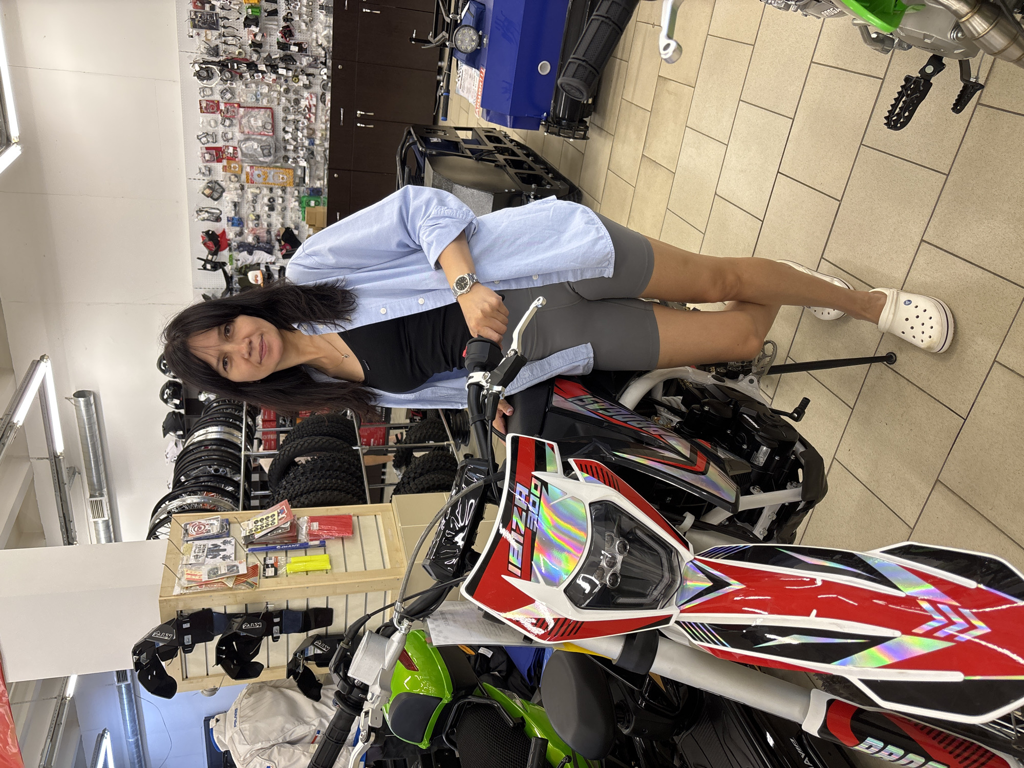 In an indoor retail environment in Chaikovsky, Russia, a woman stands next to a dirt bike. She is wearing a light blue button-down shirt over a black tank top, gray biker shorts, and white Crocs. She has dark hair and is looking towards the camera. The dirt bike is predominantly red and white with holographic decals. In the background, shelves are stocked with motorcycle parts and accessories, including helmets, wheels, and tires. A green motorcycle and a blue snowmobile are also visible. The flooring is tiled.