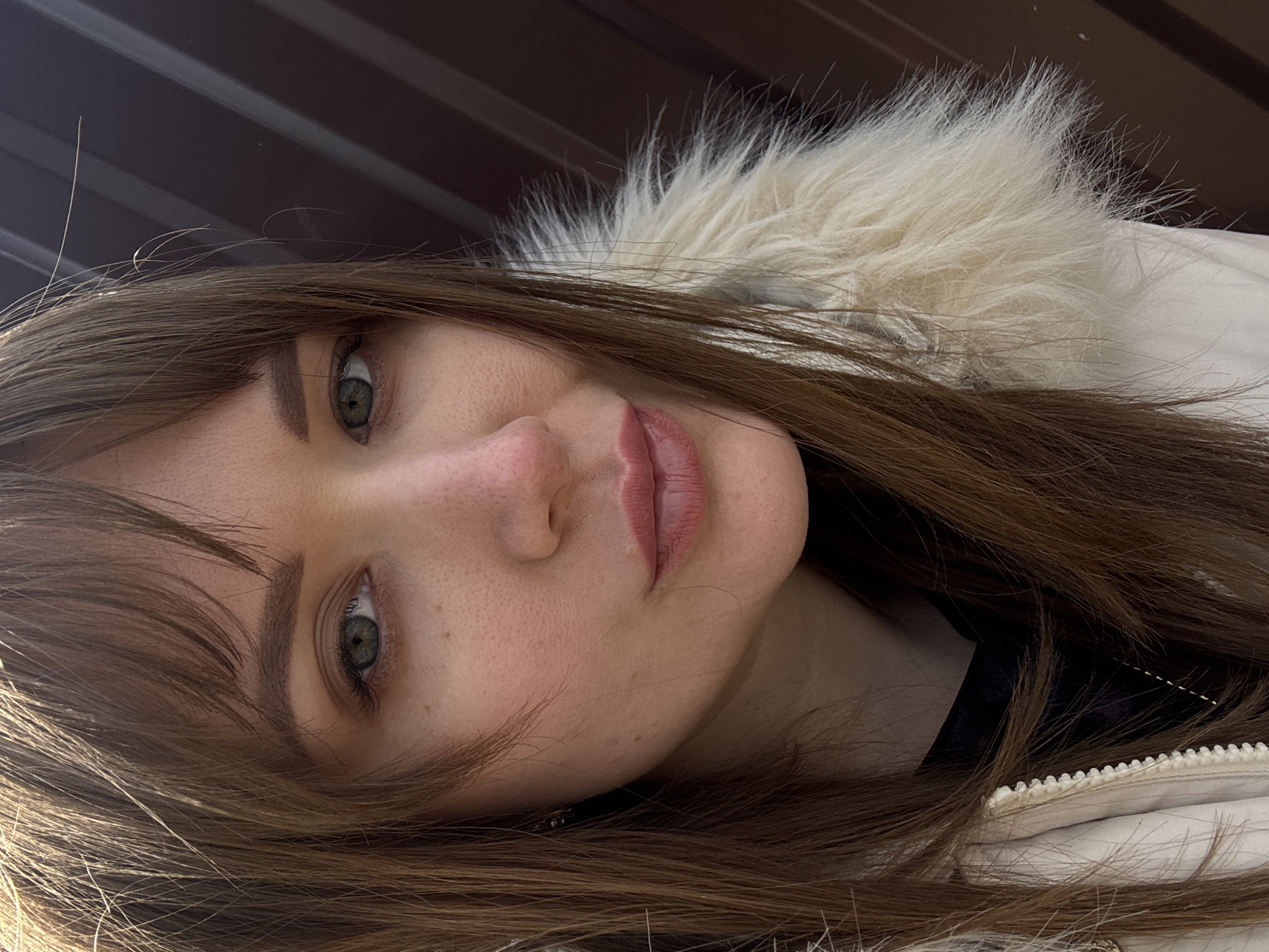 A close-up portrait of a woman with light skin, brown hair, and green eyes. She is wearing a white jacket with a fur collar. The background appears to be a dark brown surface, potentially a wall or building. There is no clear evidence to identify this image as taken in the city of Chaikovsky, Russia.