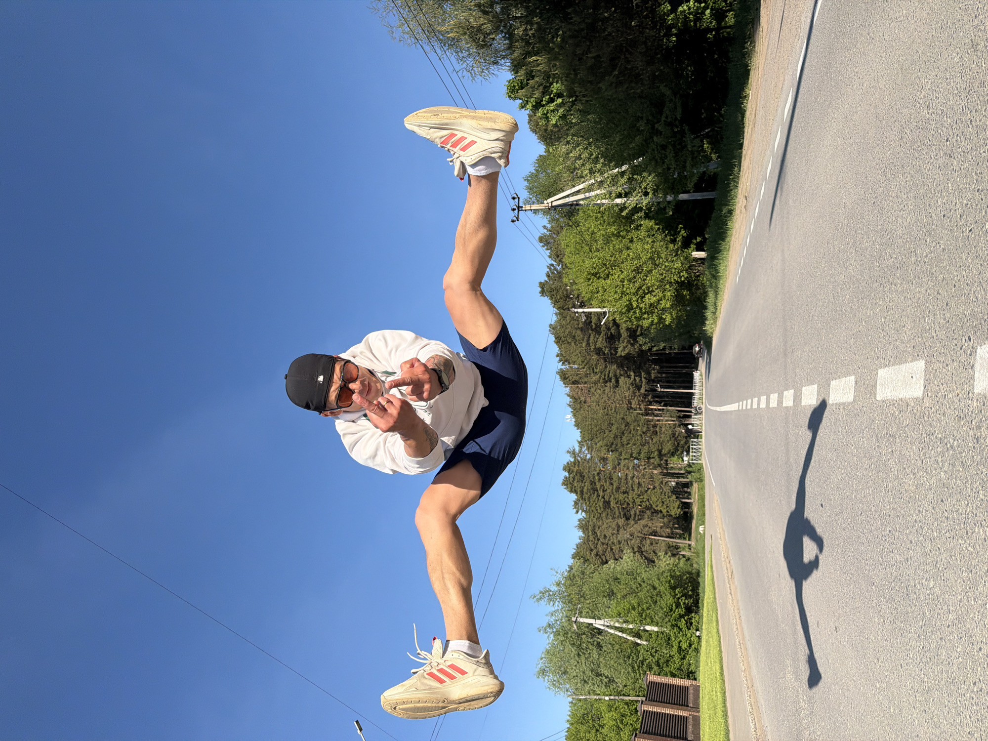 A man is posing for a photo on a road in what seems to be Chaikovsky, Russia. The road runs horizontally across the frame, with trees lining one side and the sky on the other. The man is oriented upside down in the frame. He is wearing a black cap, sunglasses, a white sweatshirt, and dark shorts. He is wearing white sneakers with red stripes. He is giving the middle finger. The shadow of the man is cast on the road.