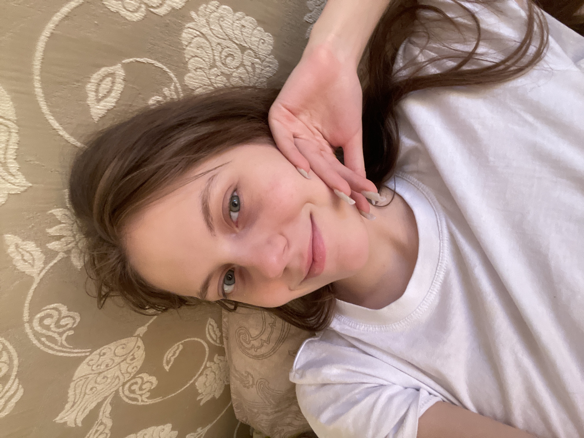 A young woman lies on a patterned, beige surface, possibly a couch or bed, wearing a white T-shirt. She is looking at the camera with a slight smile, her hand gently touching her face. The scene is likely indoors, given the lighting and background. There is no explicit information suggesting the location is Ufa, Russia.