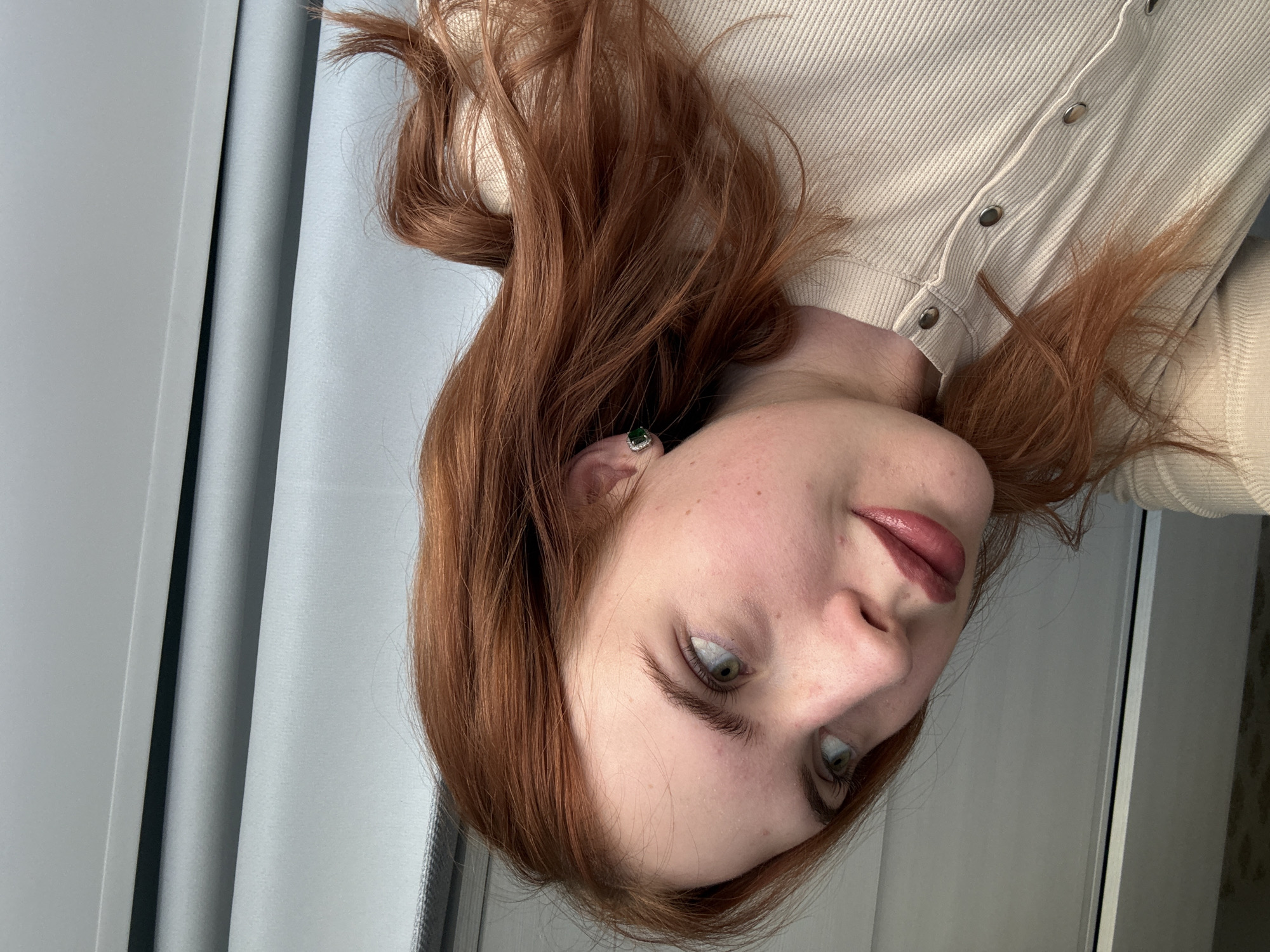 This media file presents a close-up portrait of a young woman, originally oriented sideways but depicting her upright. She has prominent reddish-brown, wavy hair that frames her face. Her fair skin features visible freckles across her nose and cheeks. She possesses green or hazel eyes, which are cast to her left, conveying a subtle and perhaps contemplative expression. Her lips are slightly parted, adorned with a rosy or berry-toned lipstick.
The woman is wearing a light-colored, possibly cream or beige, ribbed top or sweater, featuring a row of silver-toned buttons down the front. A small, square-shaped earring is visible in her left ear, characterized by a striking green gemstone (likely an emerald) encircled by tiny white stones. The background consists of soft, light grey or light blue curtains, indicating an indoor setting, possibly near a window, with natural daylight providing gentle illumination. The overall mood of the image is intimate and tranquil. This scene is located in Chaikovsky, Russia. No text is visible within the image.