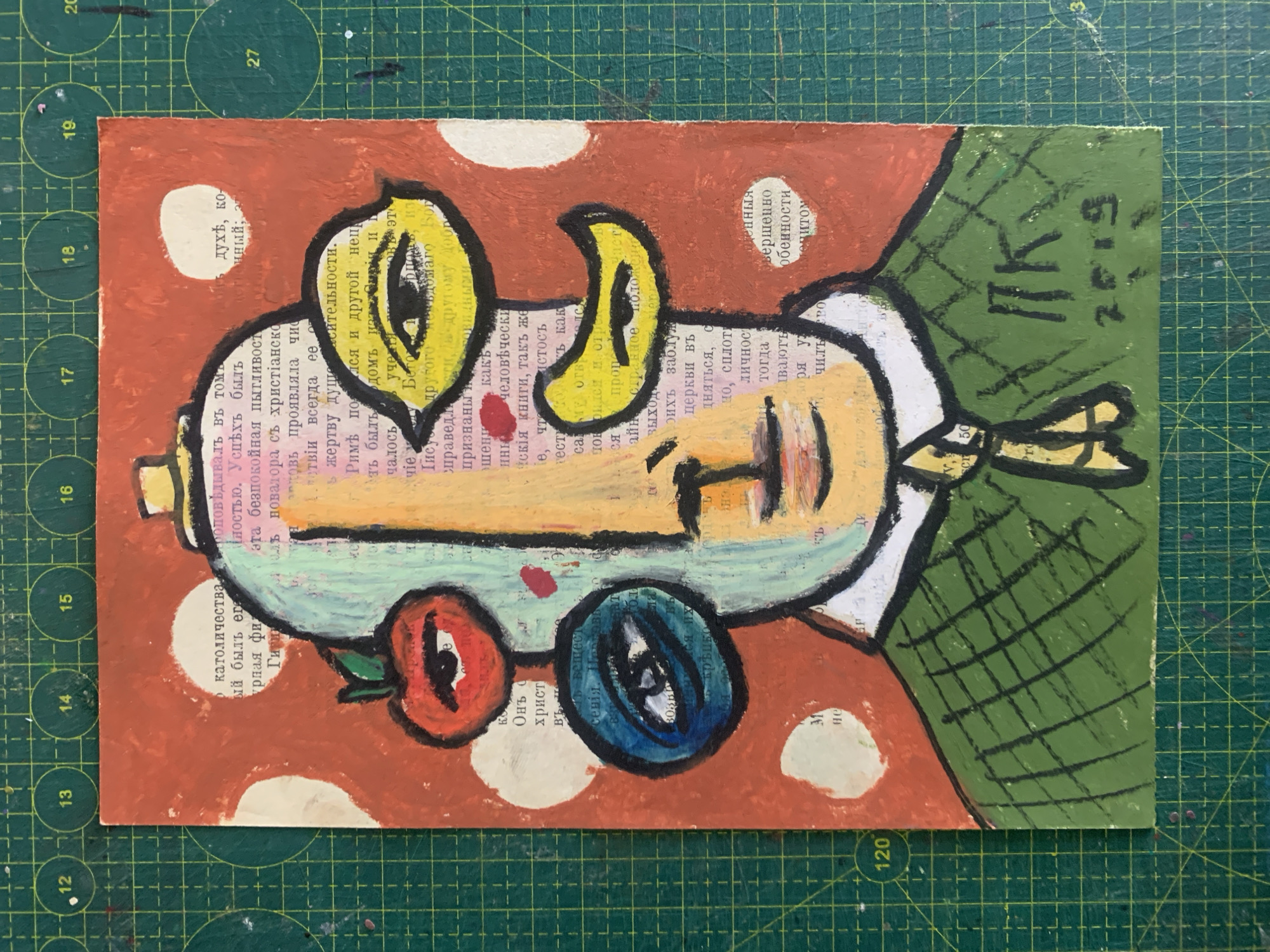 The image displays a colorful, abstract portrait painted on a collage of vintage book pages. The portrait depicts a face with multiple eyes and exaggerated features, including a red apple where one eye would typically be. The face is framed by a light blue shape, and the background features a pattern of red and white circles. The subject is wearing a green, checkered jacket. The artwork appears to be a mixed-media piece, combining painting and collage techniques. The bottom right corner of the artwork shows a signature and a date, "ПК 2019". The painting is placed on a green cutting mat with grid lines, suggesting it is in an art studio or workspace. There is no discernible location in the city of Sergeevo, Russia, depicted in this image.