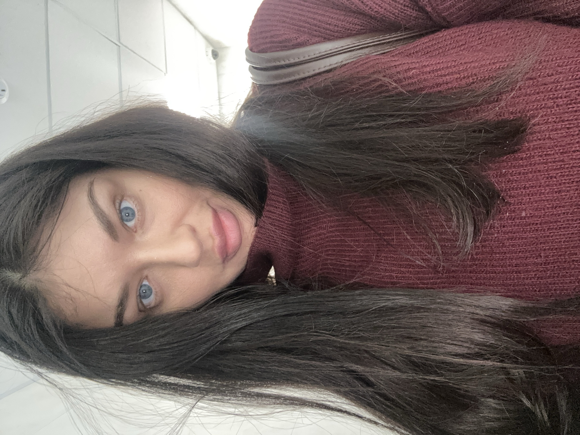 A close-up, rotated portrait features a young woman with long, dark hair and striking blue eyes. She is wearing a dark red or maroon knitted turtleneck sweater, with a brown strap, likely from a bag, visible over her shoulder. Her lips are slightly parted in what appears to be a gentle, engaging smile or playful expression as she looks directly at the camera. The background is a brightly lit, plain white wall, suggesting an indoor setting, possibly a modern building or corridor in Khimki, Russia. Vague ceiling fixtures are visible in the upper corners. The lighting is bright and even, indicating daytime indoors. No text is visible in the image.