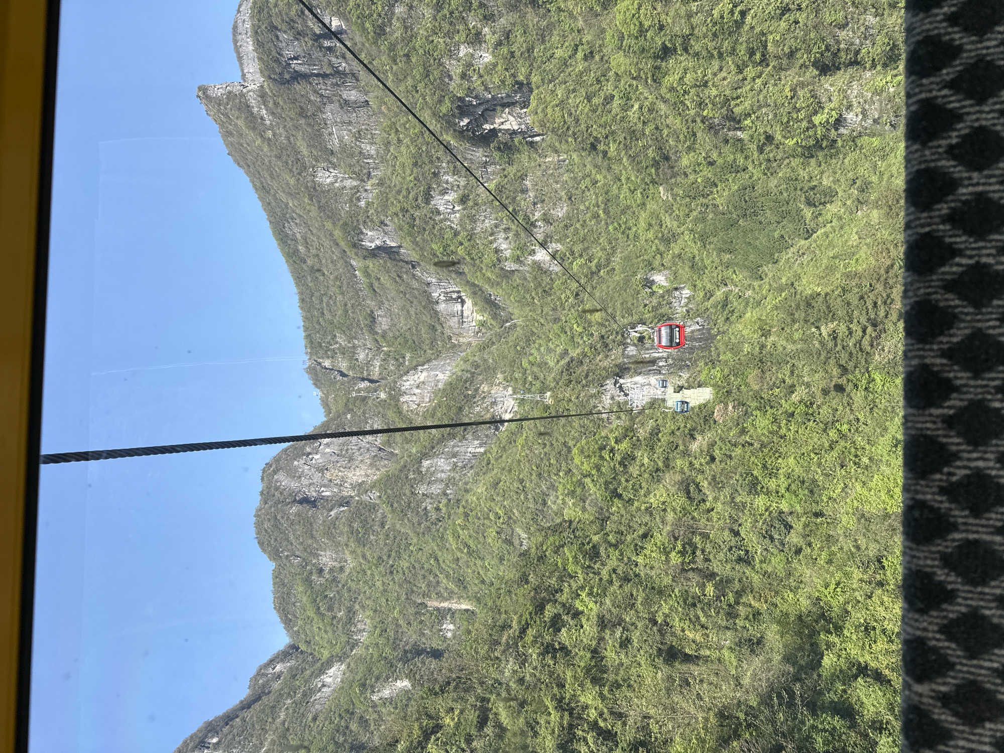 The media file presents a scenic view of a mountainous landscape, captured from inside a cable car. The perspective shows a yellow frame of the cable car window on the left side, and a dark patterned fabric from the interior on the right side, framing the expansive outdoor scene.
The dominant subjects are towering, steep mountains and cliffs, heavily covered in lush green vegetation, suggesting a vibrant natural environment. Rocky outcrops and exposed grey rock faces are interspersed among the foliage. A clear, bright blue sky fills the upper left portion of the image.
Multiple thick, dark cables of a cable car system traverse the scene diagonally. In the mid-ground, a striking red cable car cabin is visible, suspended in the air along one of these cables. Further into the distance, higher up on the mountain, several light-colored cable car cabins and their support towers can be discerned, indicating an extensive transportation network.
The activity taking place is a journey by cable car over this dramatic terrain, likely offering tourists panoramic views. The bright natural lighting and clear blue sky suggest it is daytime, possibly a sunny morning or afternoon. The scene evokes a sense of awe and adventure. There is no visible text in the image. This landscape is characteristic of the impressive natural scenery found in regions like Yongding District, China, known for its karst formations and deep valleys.