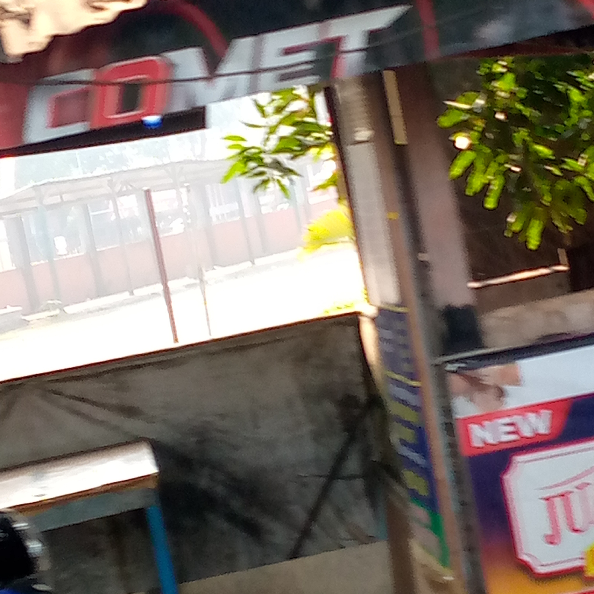 The image depicts an outdoor scene in Tambakagung, Indonesia, likely near a food or beverage stall. In the foreground, a weathered, greyish-blue tarp is visible. To the right, a sign for "NEW JUICE" is prominently displayed in red and white lettering. Behind this, a bright green leafy plant adds a touch of nature to the scene. Further back, a light-colored structure with a red roof and white poles suggests a covered seating or serving area. The overall environment appears to be urban or semi-urban, with hints of commercial activity.