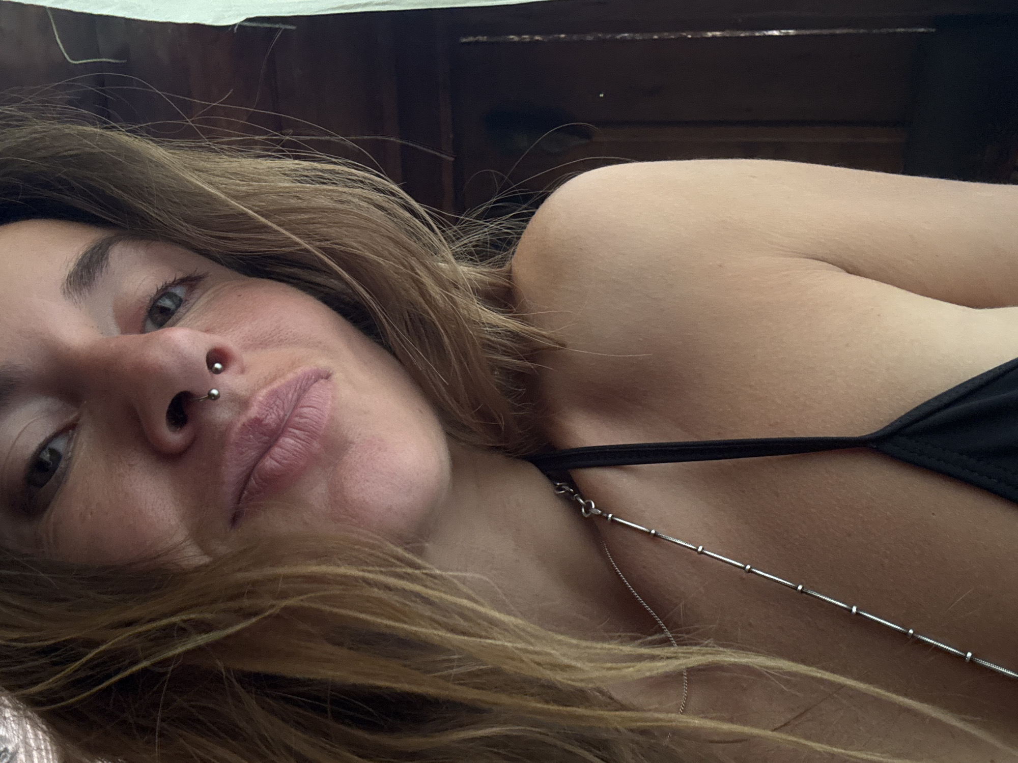 A close-up, head-and-shoulders shot captures a young woman with blonde hair and a septum piercing. She is wearing a black bikini top and a silver necklace. The background is dark and appears to be an indoor setting with wooden elements. Her head is tilted slightly, and her gaze is directed towards the camera. The lighting is soft, highlighting her facial features and skin texture.