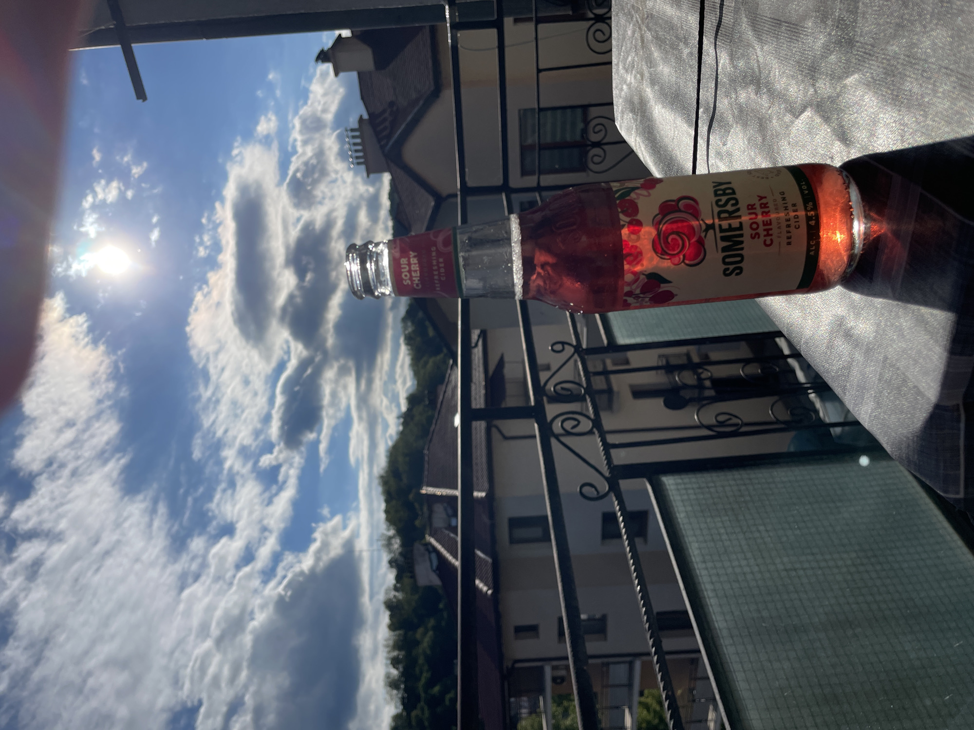 The image depicts a bottle of Somersby Sour Cherry cider lying horizontally on a surface, likely a table, with the label facing the viewer. The bottle is partially in sunlight. In the background, there's a view through what appears to be a balcony railing. The sky is visible, filled with numerous white clouds against a blue backdrop. Buildings and trees can also be seen in the background.