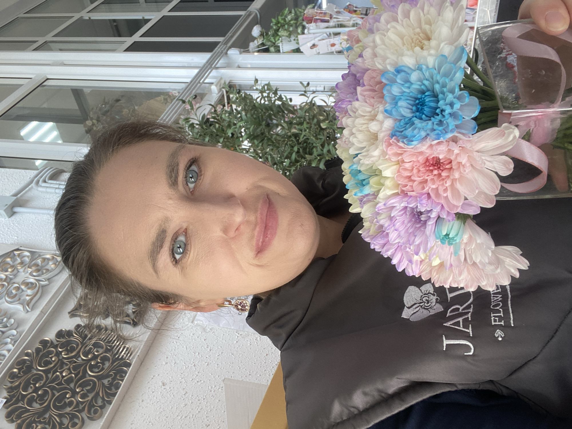 A woman is holding a bouquet of colorful chrysanthemums. The flowers are in shades of white, blue, pink, and purple. The bouquet is contained in a clear glass vase tied with a light pink ribbon. The woman is wearing a black jacket with white lettering that reads "JAR . FLOW" and includes an image of a flower. She has light skin, blue eyes, and her hair is pulled back. She is wearing stud earrings. The background includes a window with a white frame, green plants, and a decorative white wall panel with a bronze-colored ornate design. The photograph was taken in the city of Yekaterinburg, Russia.