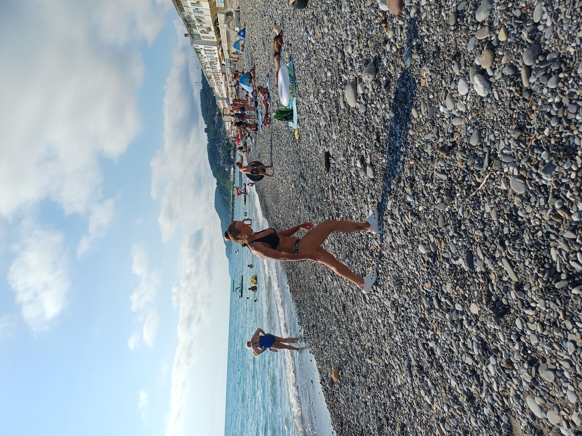 A wide shot of a crowded pebble beach in Sochi, Russia, under a partly cloudy sky. The sea is calm with small waves lapping the shore. In the foreground, a woman in a black bikini and white Crocs walks towards the viewer. Behind her, several other people are scattered across the beach, some sunbathing, others standing by the water. Buildings are visible on a hillside in the background, lining the coast. The scene is typical of a sunny day at a Russian Black Sea resort.