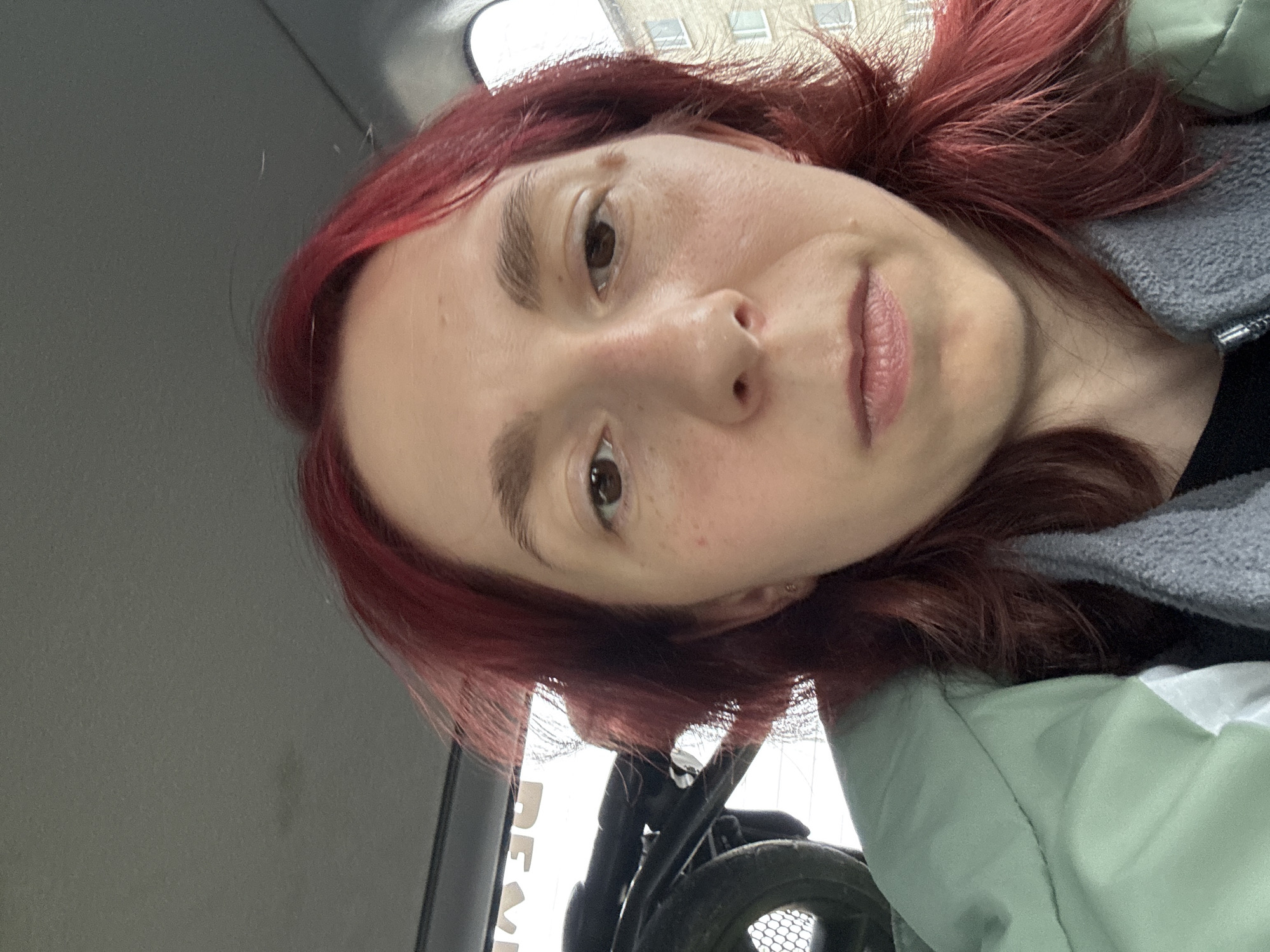 A close-up, selfie-style photograph of a woman with bright red hair, facing the camera. She is inside what appears to be a vehicle, with a dark gray headliner visible in the upper portion of the frame. A portion of a dark-colored object, possibly a stroller or luggage, with a wheel, is visible on the left side of the image. The woman is wearing a light green jacket over a gray scarf or hooded garment. The background outside the vehicle shows a building and some trees. The lighting suggests it is daytime. The location is identified as Unknown, Unknown.