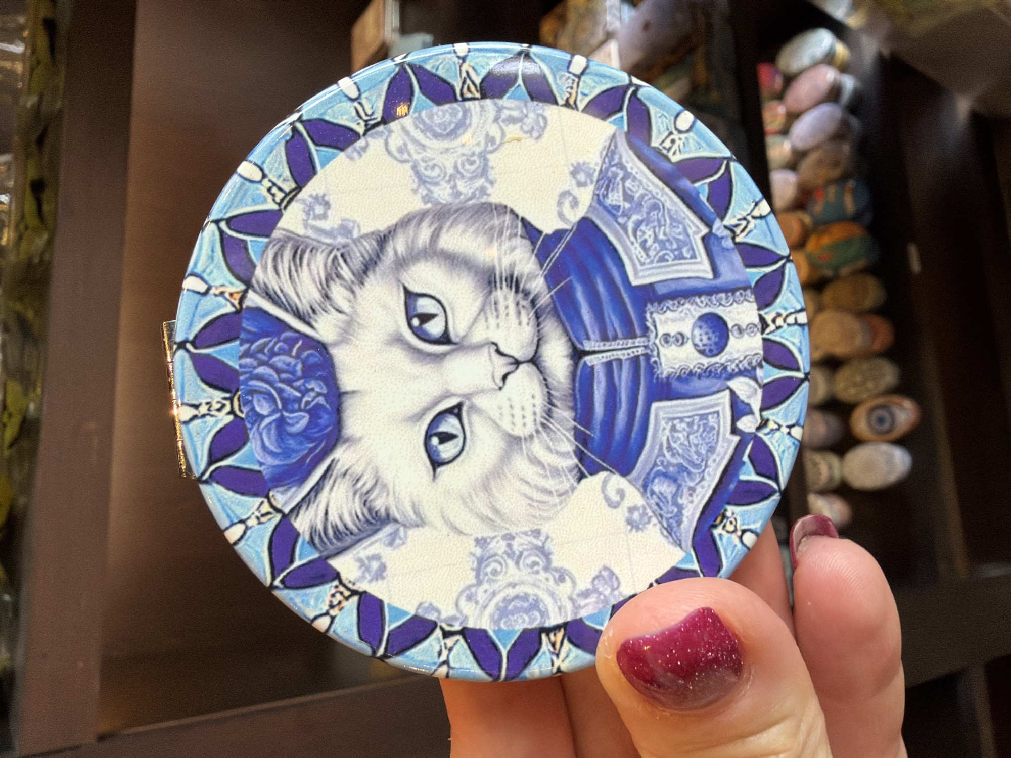 A circular compact mirror featuring a stylized portrait of a white cat wearing a blue hat and jacket is held by a person's hand with dark pink nail polish. The cat has blue eyes and is depicted against a background with intricate blue and white patterns. The mirror has a metallic gold-colored edge. The background appears to be a shelf or display, with out-of-focus items visible below, including small decorative objects. The image is taken indoors. The person's thumb and index finger are visible holding the mirror, with the fingernail of the index finger prominent.